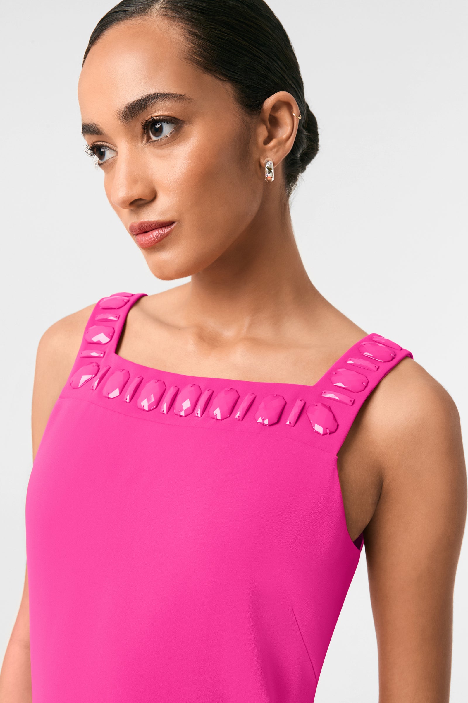 A woman in the Yokuna Jewel Neck Mini Dress, a bright pink sleeveless style with glossy accents at the square neckline, wears small hoop earrings and looks slightly to the side against a plain light background.