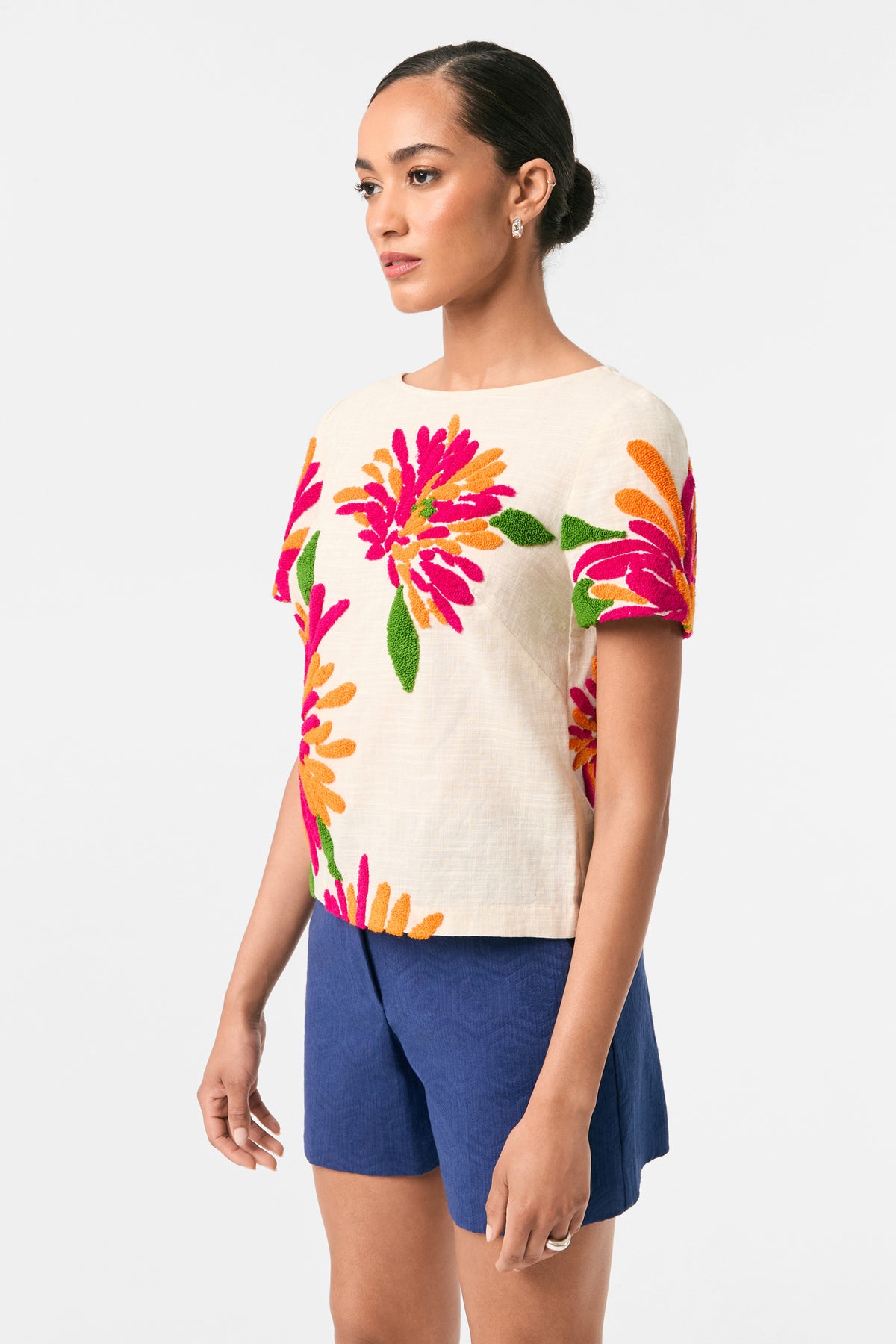 A woman stands against a white background wearing the Siona Embroidered Flowers Top in cream with pink, orange, and green floral embroidery, paired with blue shorts. Her hair is neatly pulled back as she gazes slightly to the side.