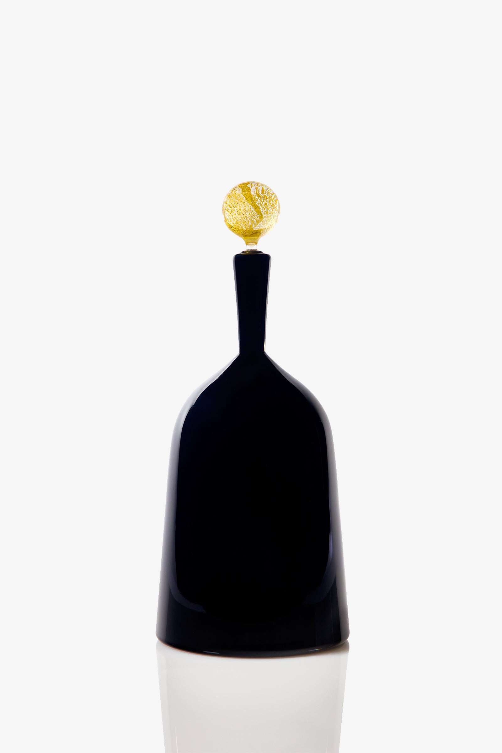 The Wide Bottle Carmella Barware is a sleek, black, bell-shaped glass vessel with a narrow neck and a small, textured golden sphere on top, set against a plain white background.