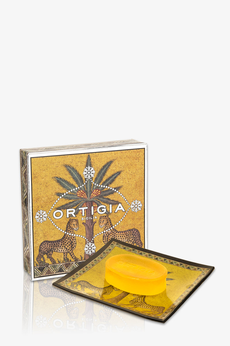 The Ortigia Glass Plate & Soap set includes a yellow soap bar on an elegant glass dish, packaged in a box with leopards, palm trees, and rich Sicilian-inspired gold and brown designs.
