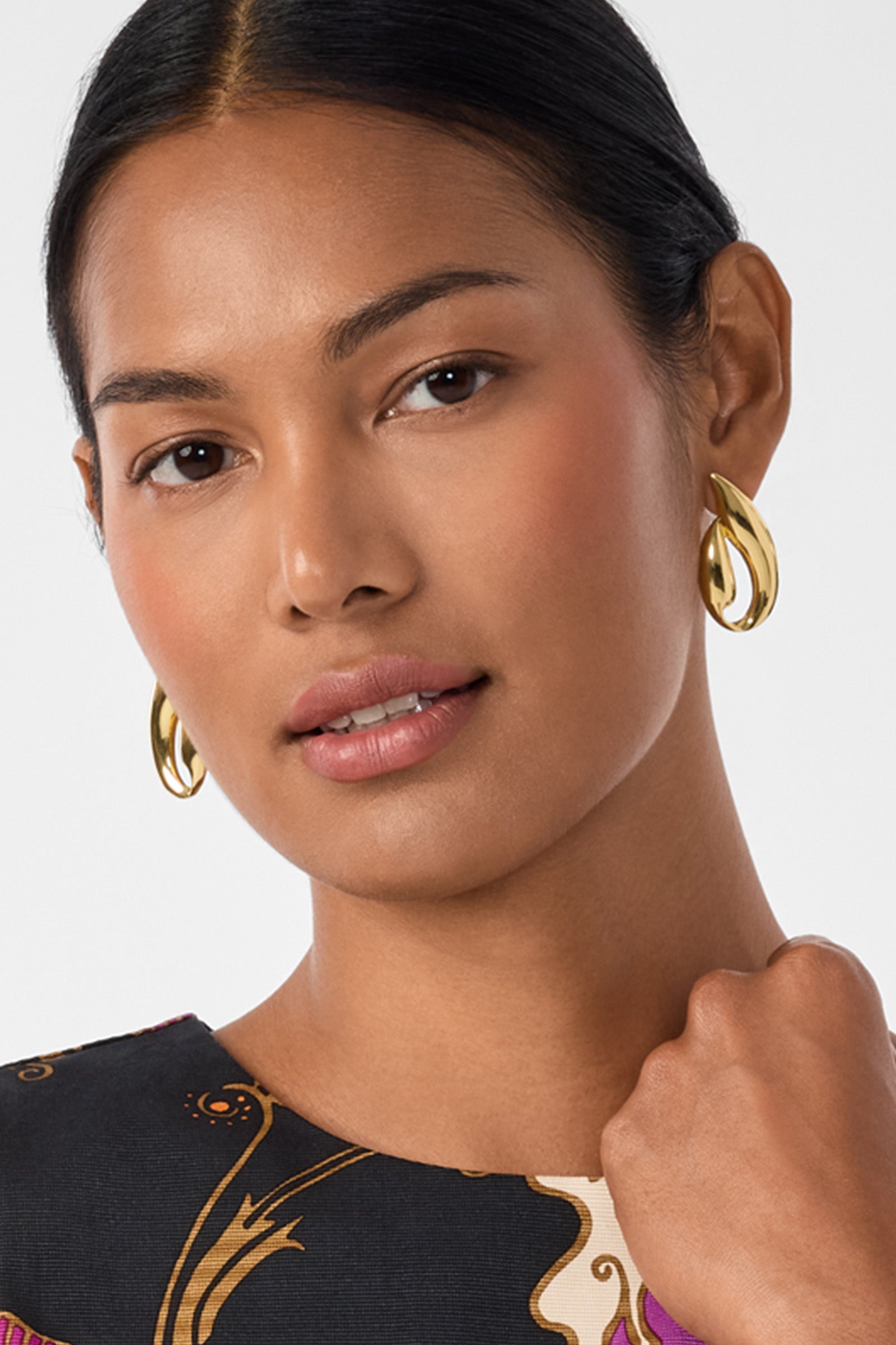 A woman with smooth, medium-brown skin and dark hair pulled back wears Abstract Post Earrings and a patterned top. She gazes confidently at the camera while gently touching her collar.