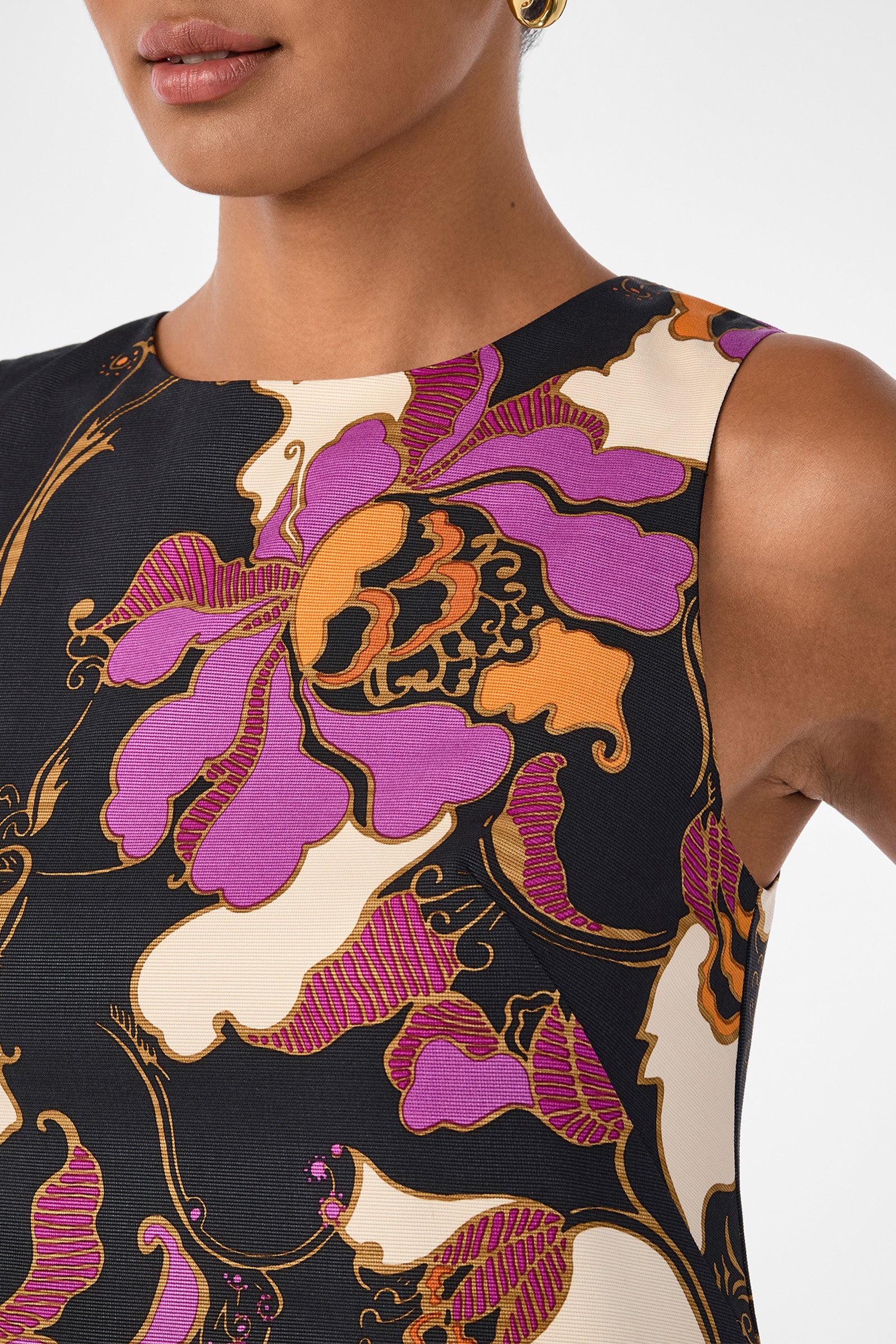 A woman wears the Addy Dress, a sleeveless piece with a bold floral pattern in purple, orange, cream, and black; only her lower face, neck, and upper torso are visible.