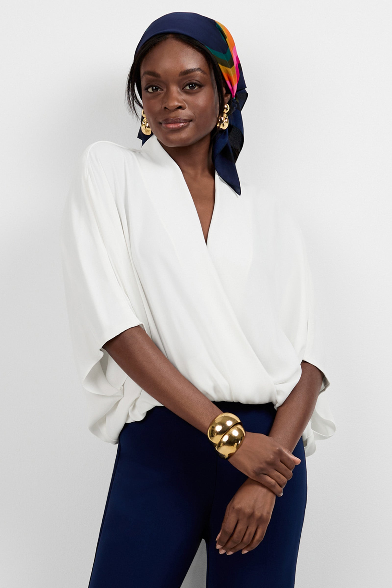A woman stands against a white wall, wearing the Anora V-Neck Wrap Top in Whitewash and navy pants. She accessorizes with a colorful headscarf, gold earrings, and a chunky bracelet, smiling softly.