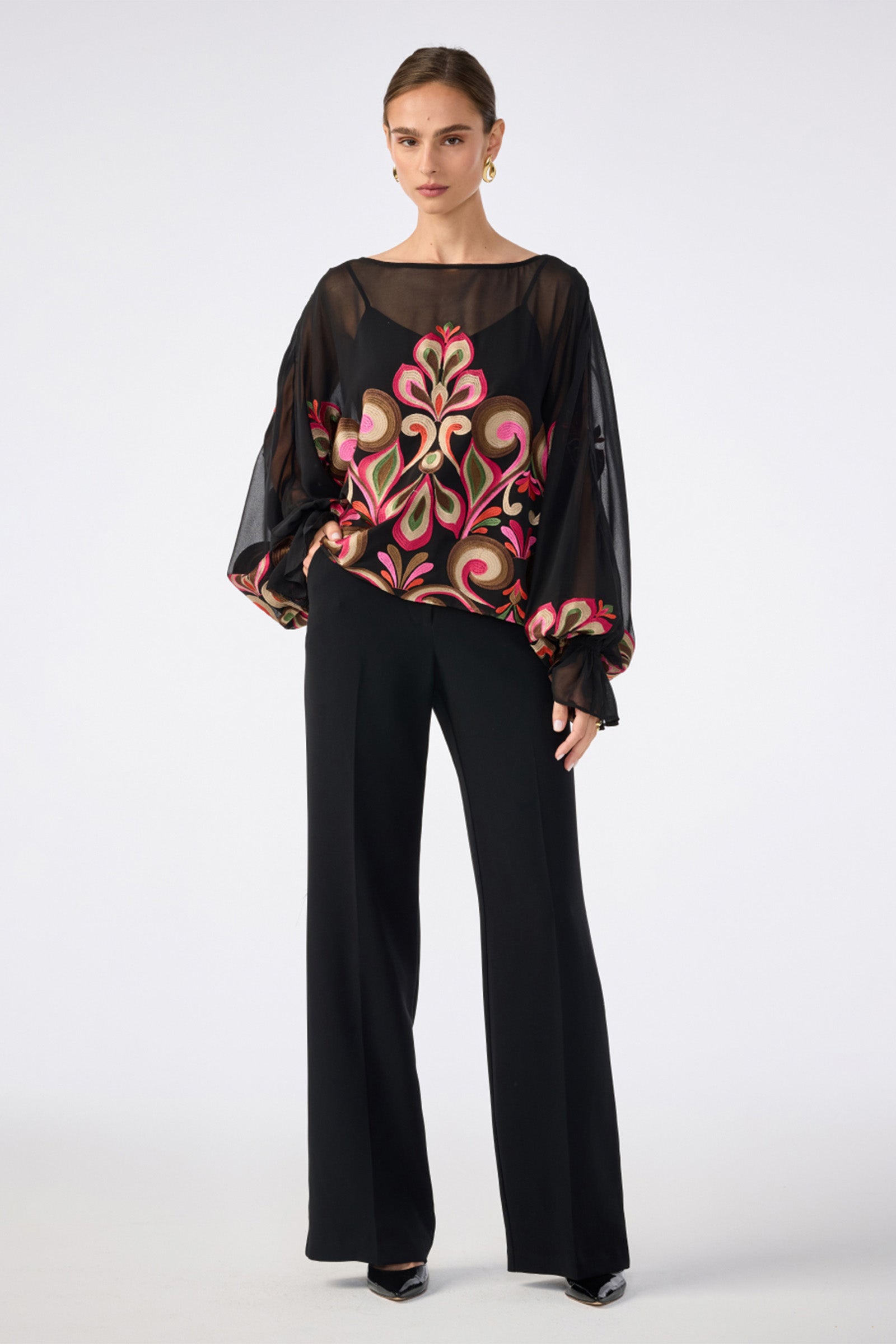 A woman wears the Soho Embroidered Top by Sanctuary—a sheer black blouse with voluminous sleeves and a bold, colorful floral front—paired with black wide-leg pants, hoop earrings, and black pointed shoes against a plain white background.