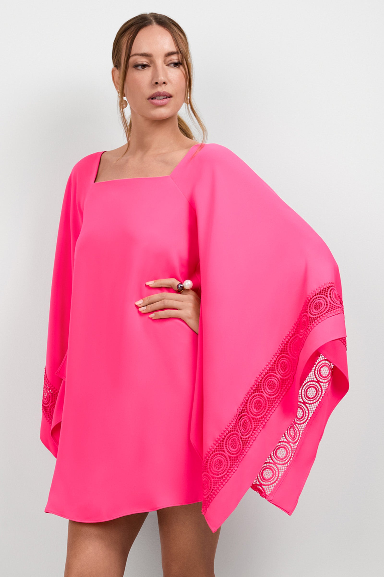 A woman models the Aqualane Side Flounce Dress, a bright pink, loose-fitting style with wide, flowing sleeves accented by circular cutouts. She has light brown hair in a low ponytail and stands before a plain light background.