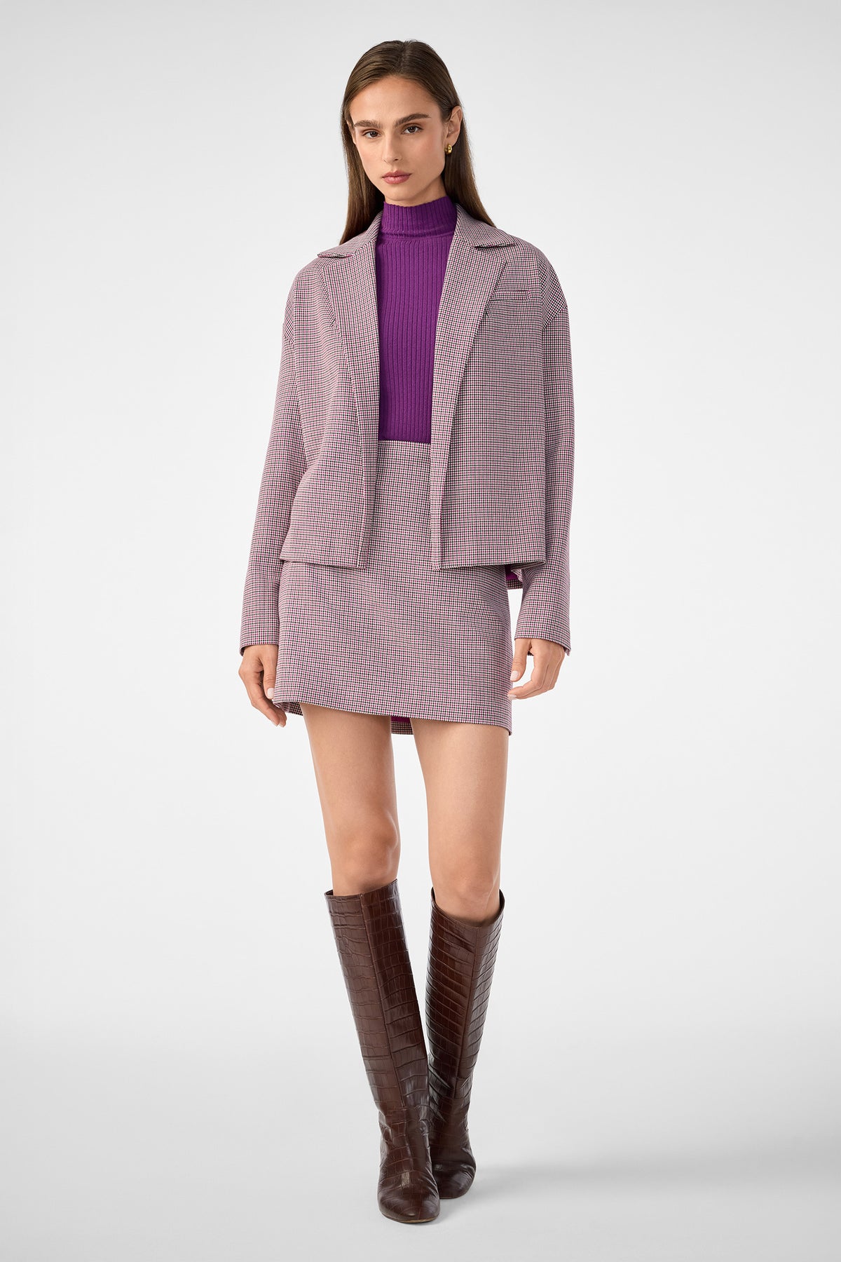 A woman stands against a plain light background, wearing the Ayla Jacket in pink and grey check with a matching mini skirt, a purple turtleneck sweater, and brown knee-high boots.