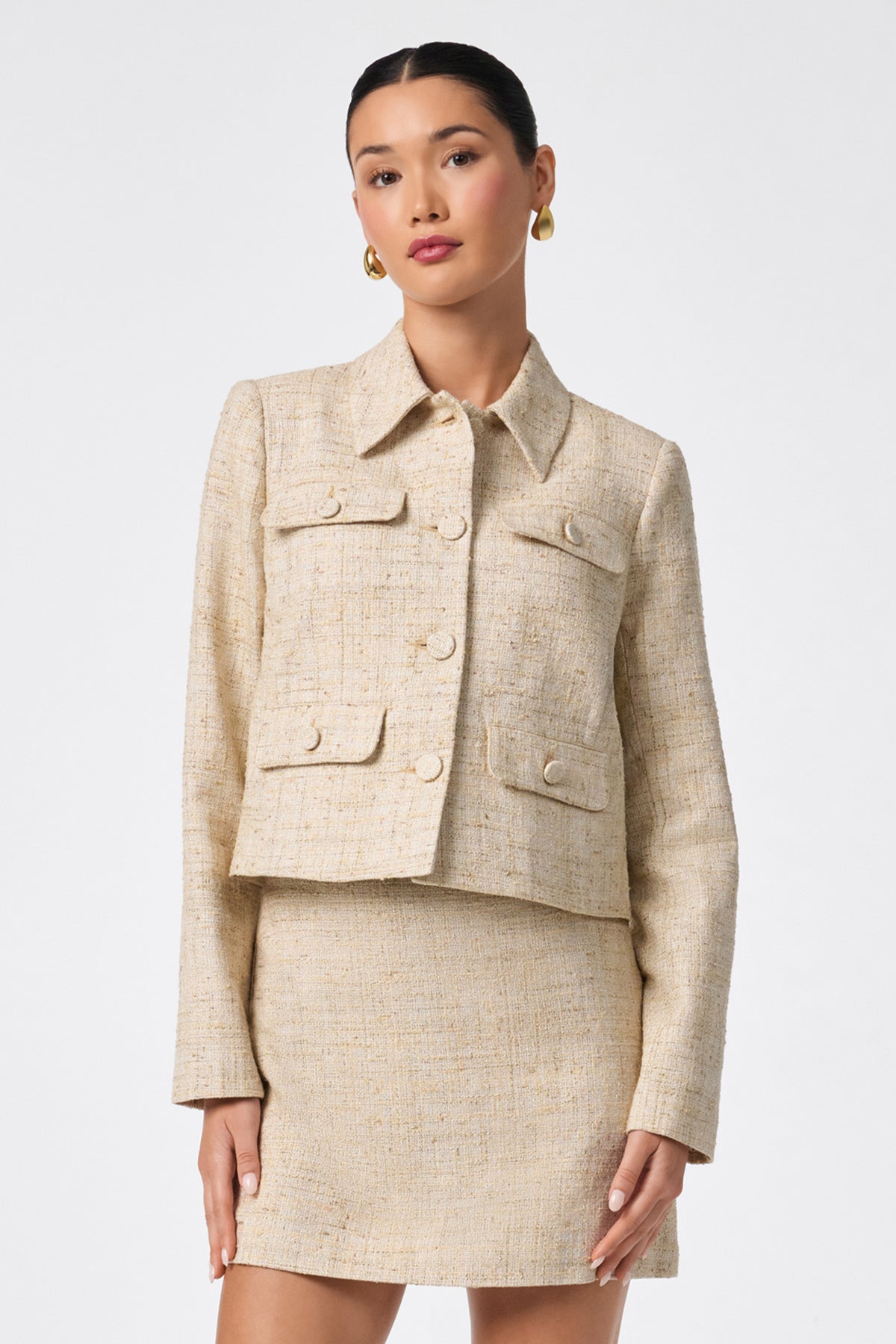 A woman stands against a white background wearing the Bayonne Jacket, a light beige tweed piece with front pockets and large buttons, paired with a matching skirt, her hair slicked back and accessorized with gold hoop earrings.