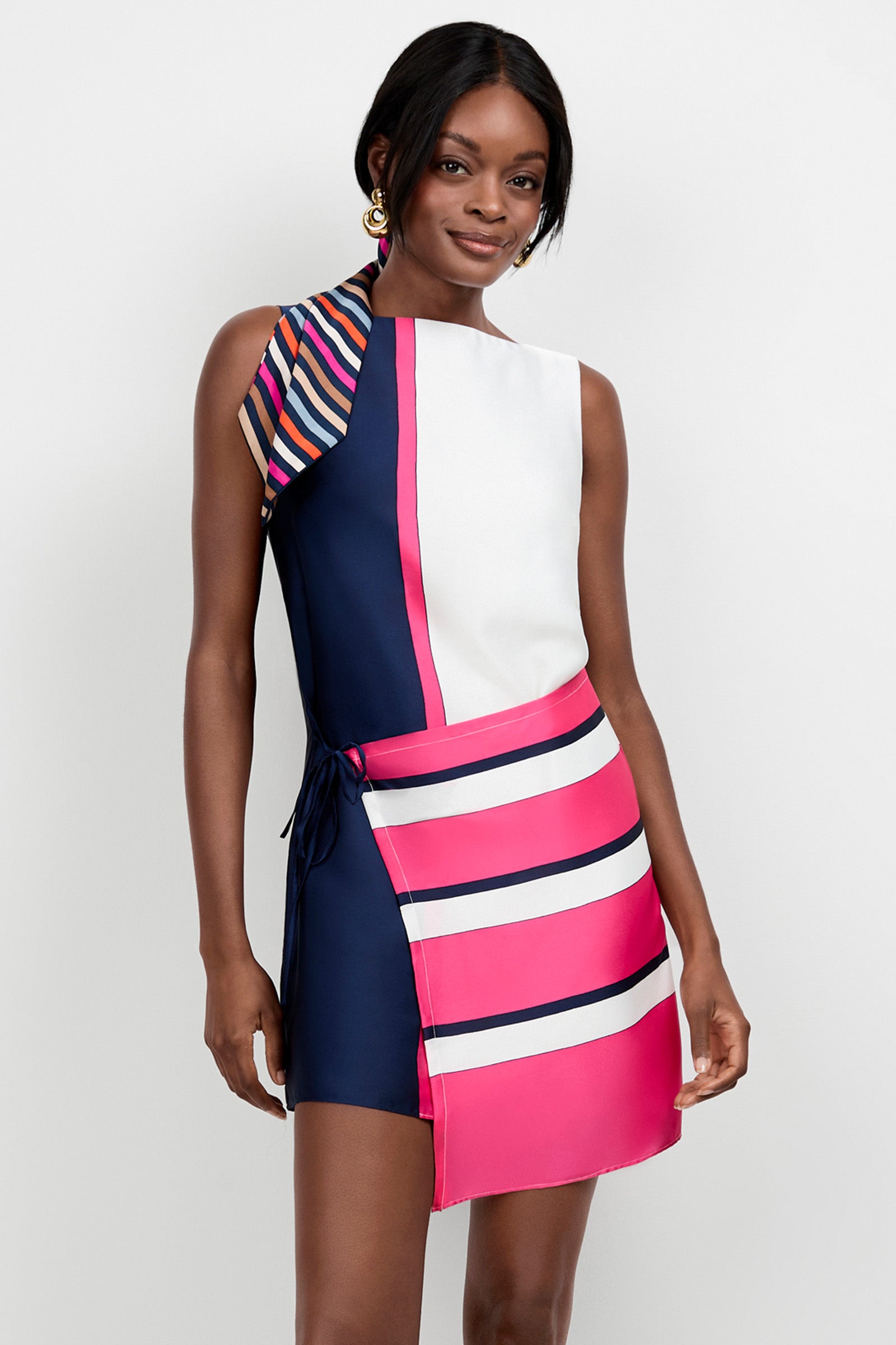 A woman wearing the Brisa Linee Mini Dress—a sleeveless, asymmetrical mini dress with bold navy, white, pink, and striped patterns—stands against a plain light background.