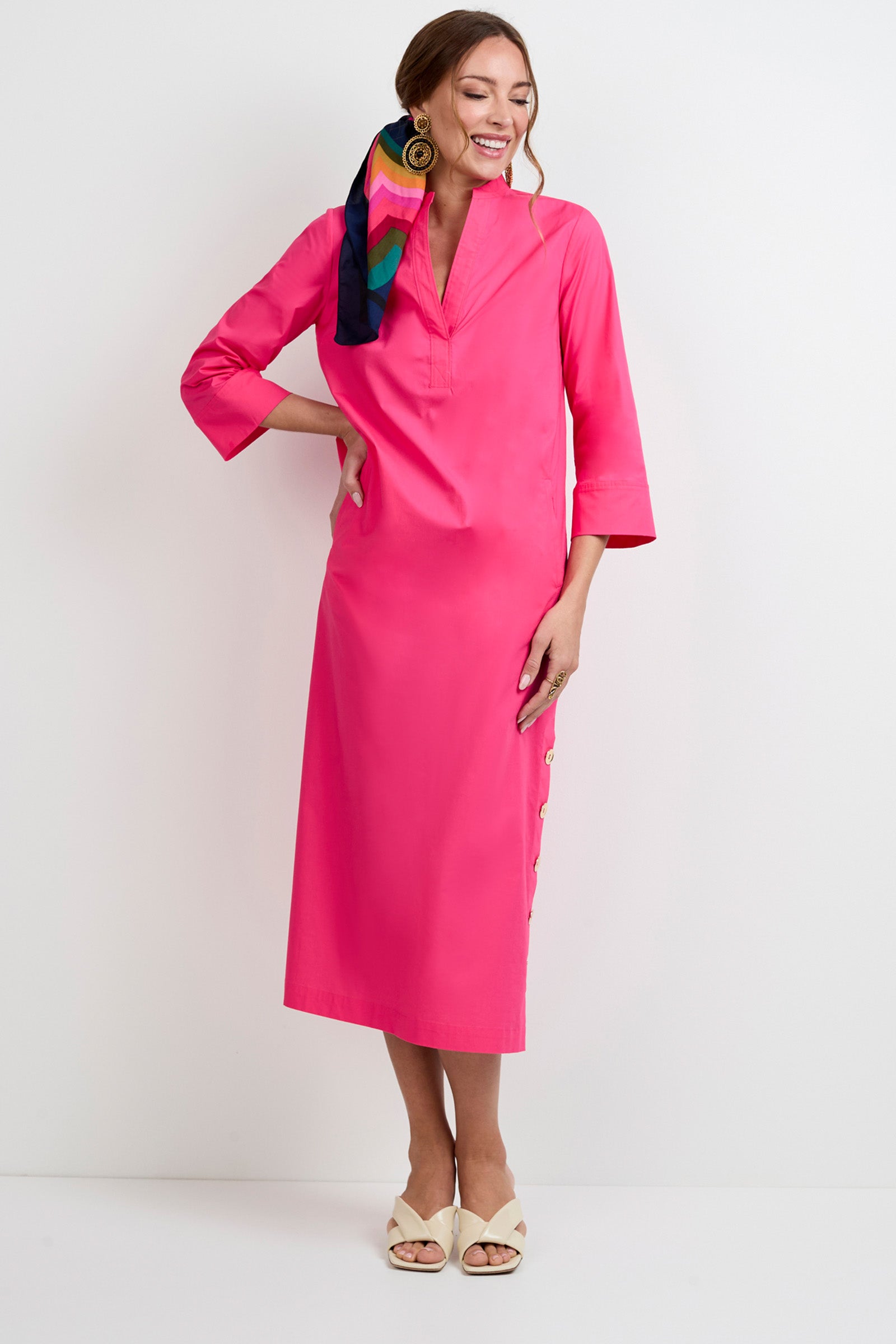 A woman smiles against a white wall, wearing the Capella Mandarin Collar Maxi Dress in bright pink, styled with a colorful hair scarf, large round earrings, and cream heeled sandals.