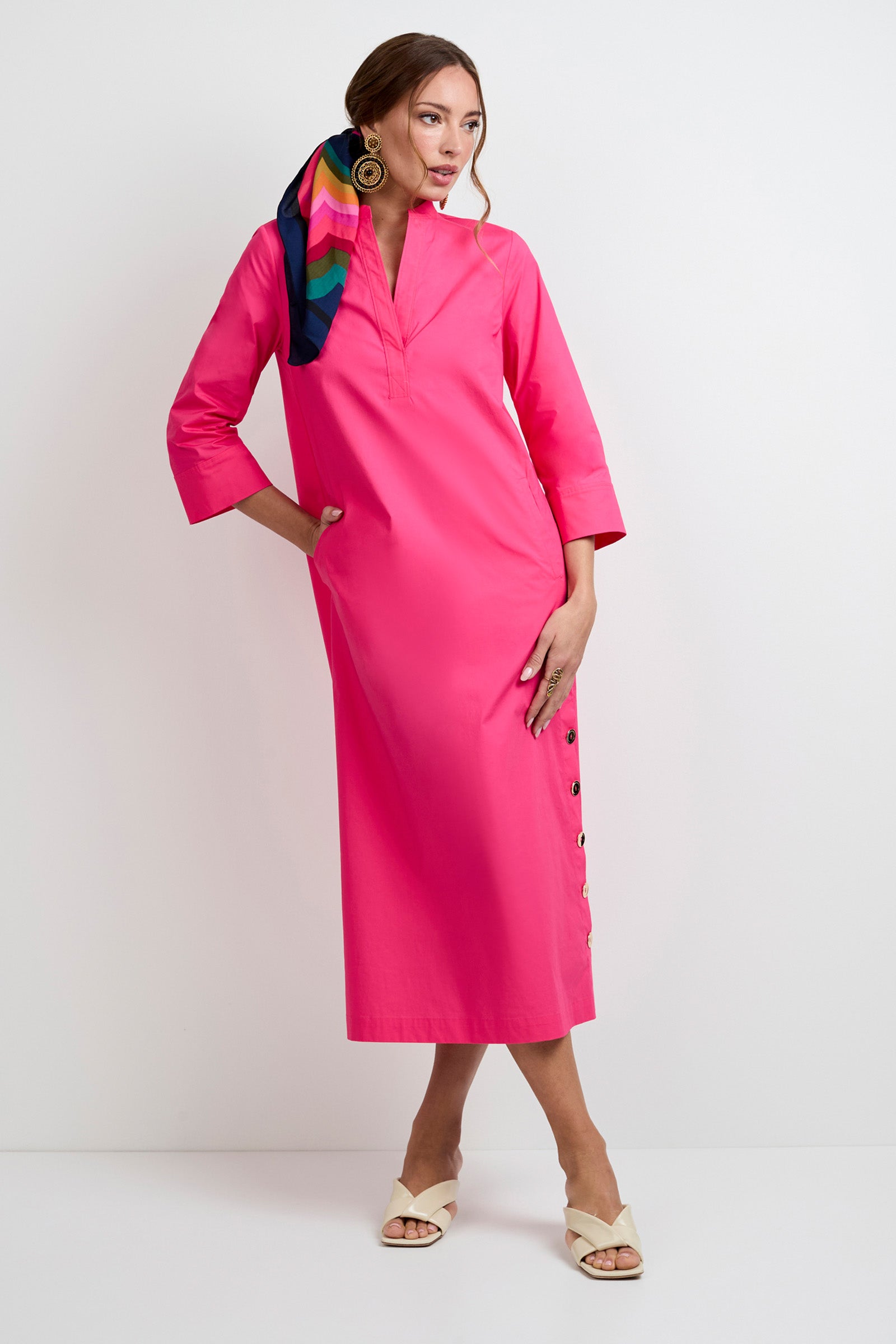 A woman poses against a plain white background wearing the Capella Mandarin Collar Maxi Dress in bright pink, paired with beige sandals, gold earrings, and a colorful scarf tied in her hair.