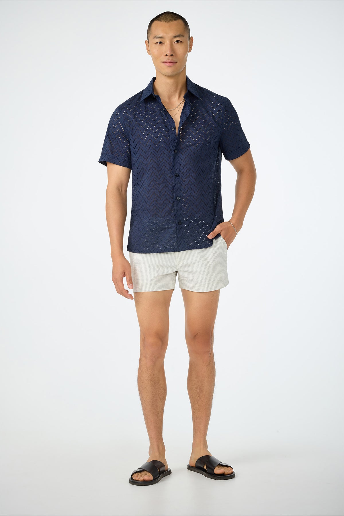 A man stands against a plain background wearing a navy zigzag shirt, Chico Swim Trunk - Seersucker white shorts, black sandals, and a gold necklace. One hand is in his pocket and he smiles slightly.