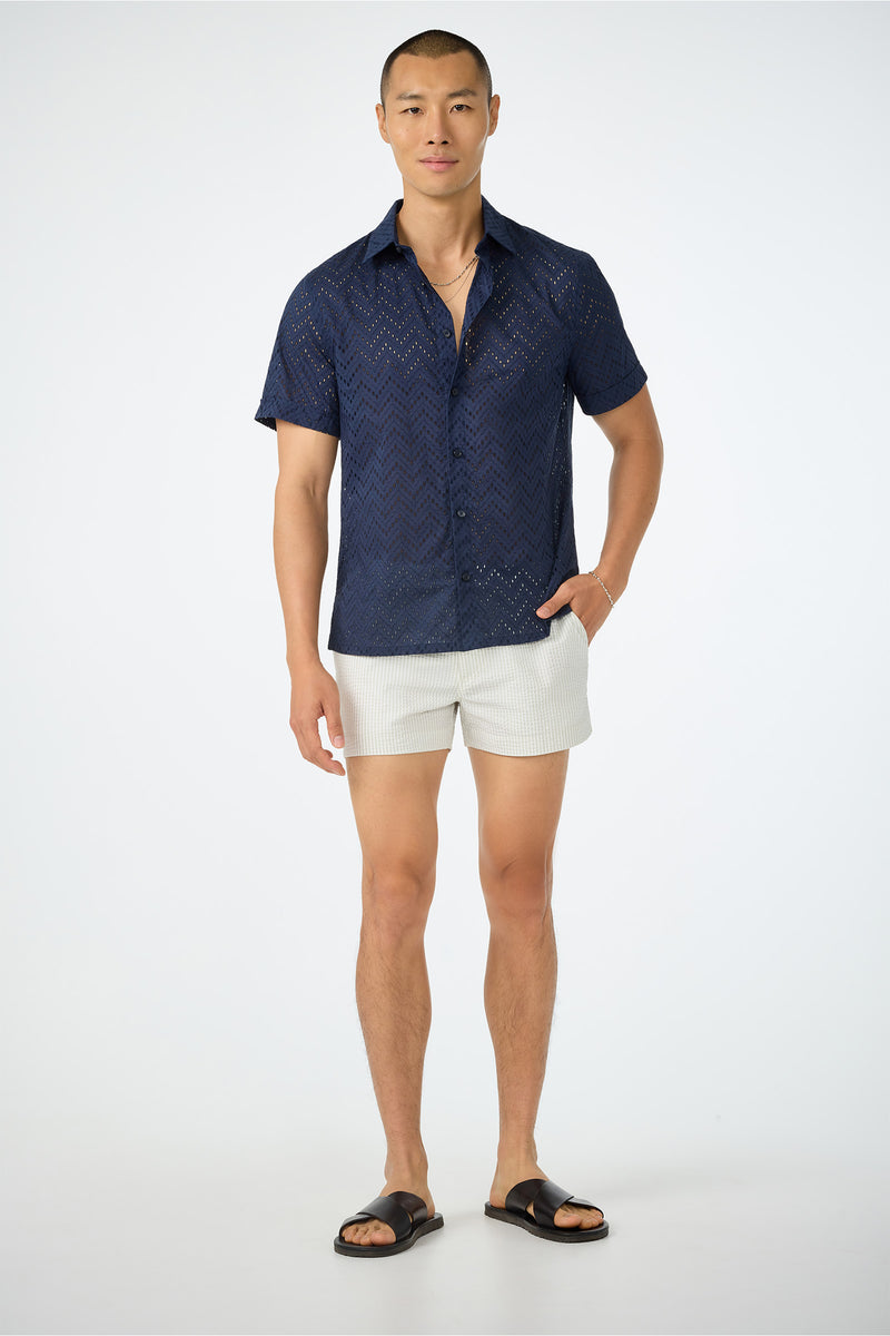 A man stands against a plain background wearing a navy zigzag shirt, Chico Swim Trunk - Seersucker white shorts, black sandals, and a gold necklace. One hand is in his pocket and he smiles slightly.
