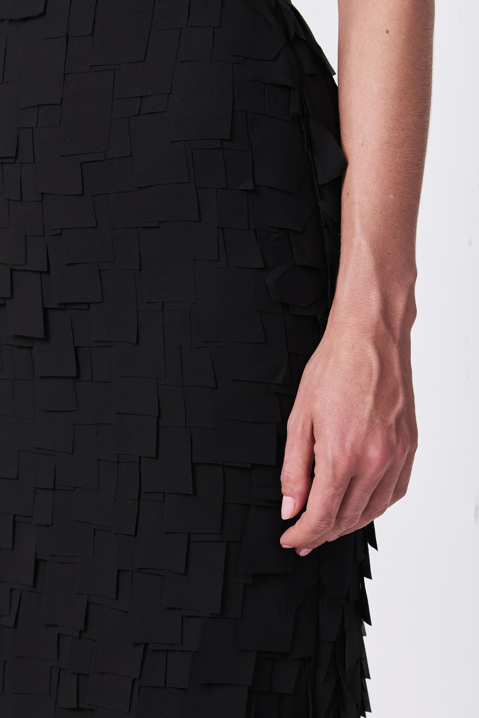 A close-up of a persons right hand resting against their thigh, wearing the Dita Midi Pencil Skirt - Flutter Chiffon, which features layered square fabric pieces for a textured geometric look.
