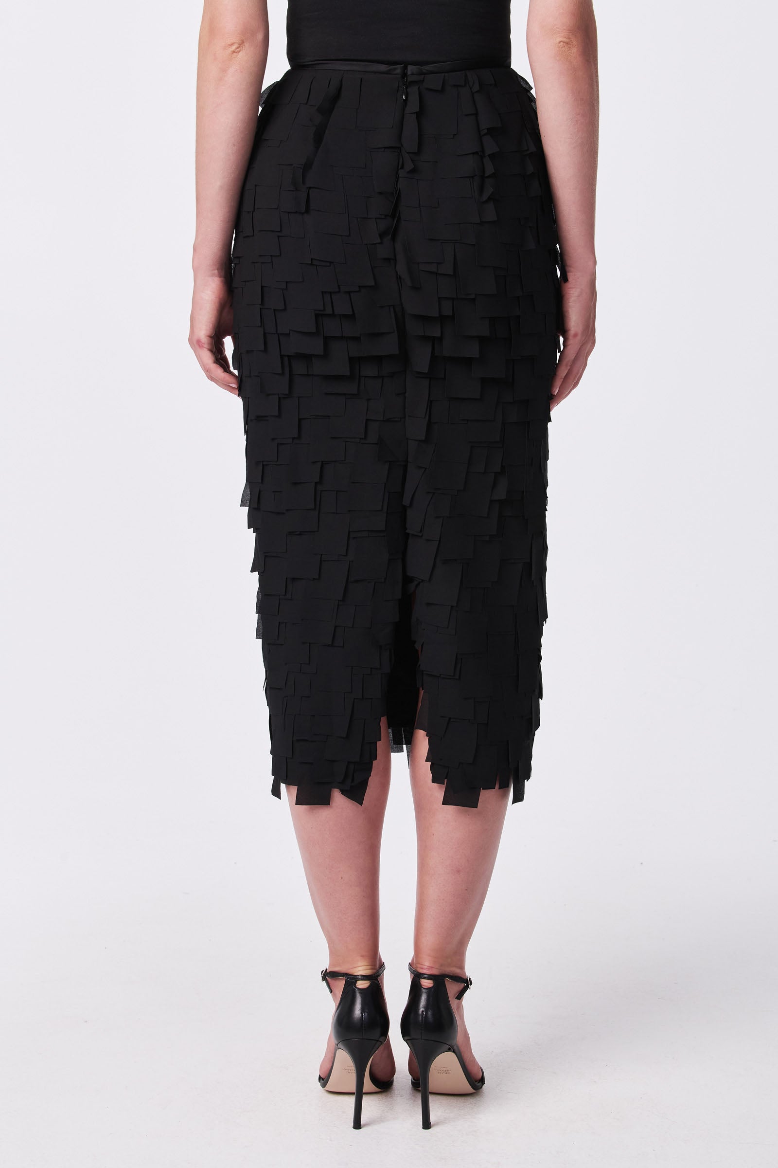 A person is seen from behind wearing the Dita Midi Pencil Skirt - Flutter Chiffon, featuring layered, square-shaped details, paired with black high-heel sandals.
