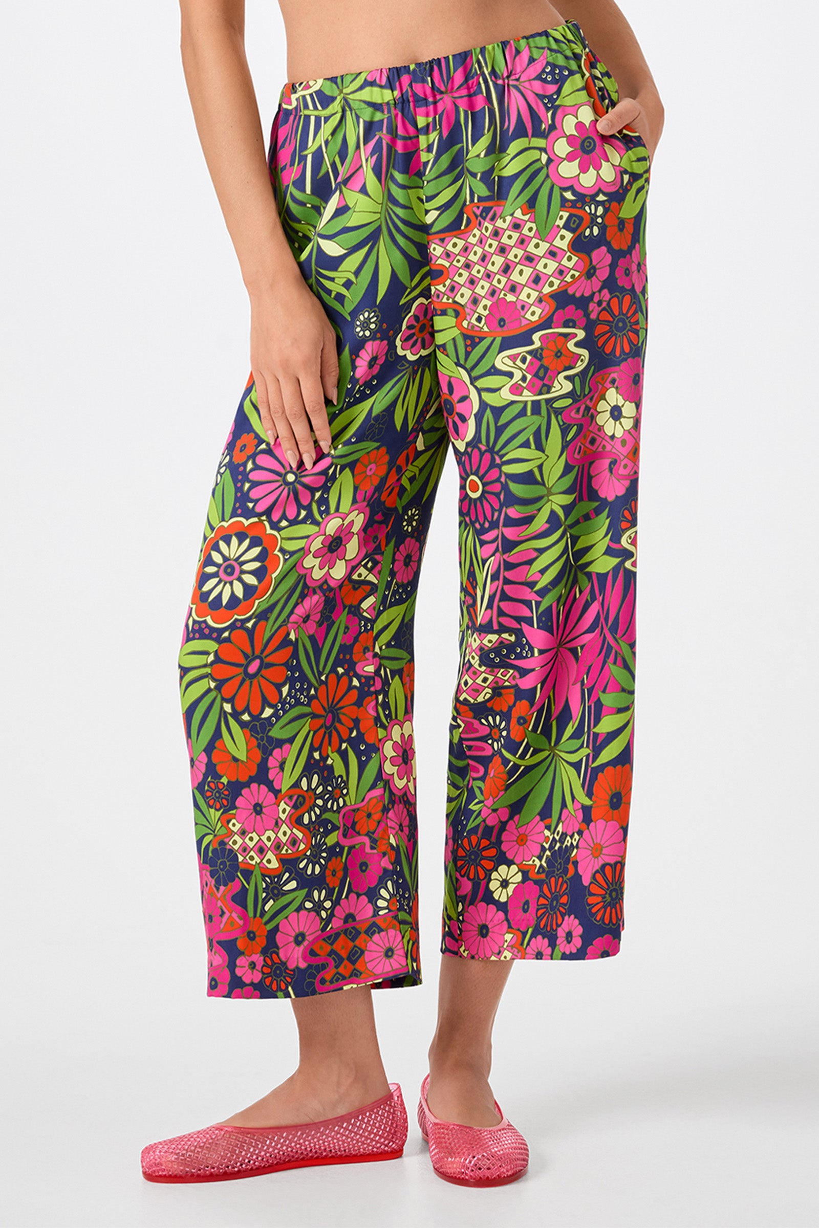 Someone wears the Estella 2 Cropped Pants - Fumi Floral, featuring a vibrant red, pink, yellow, and green floral pattern. Paired with pink flats, only their lower half is visible against a plain background.