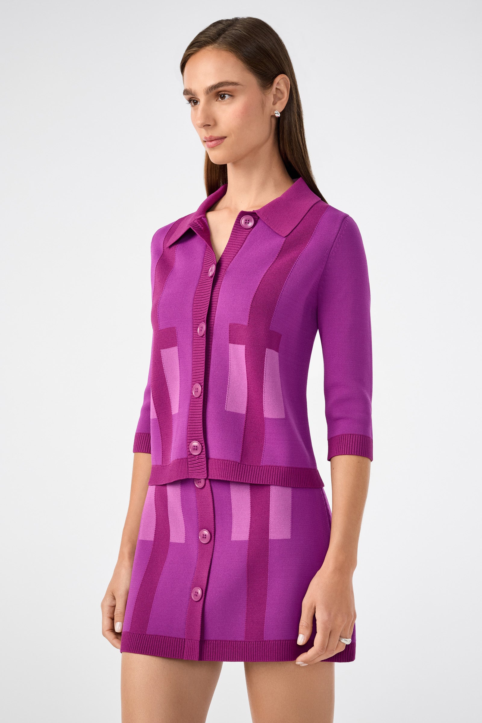 A woman wears the Gaia Cardigan, a purple button-up with half sleeves, paired with a matching high-waisted geometric skirt, standing against a plain light background.