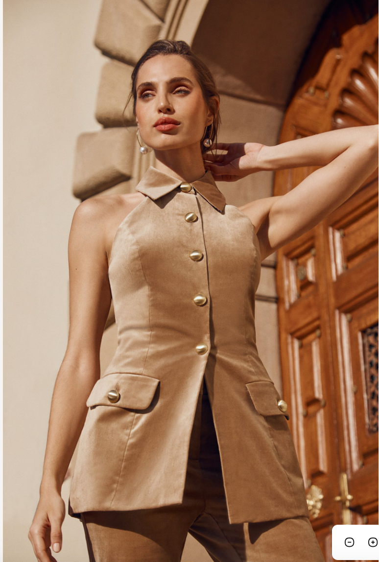 A woman poses in front of a wooden door, wearing the Elainia Velveteen Sleeveless Tunic Top in GOLD, featuring large buttons and flap pockets. With one hand raised to her forehead, she gazes confidently into the distance.