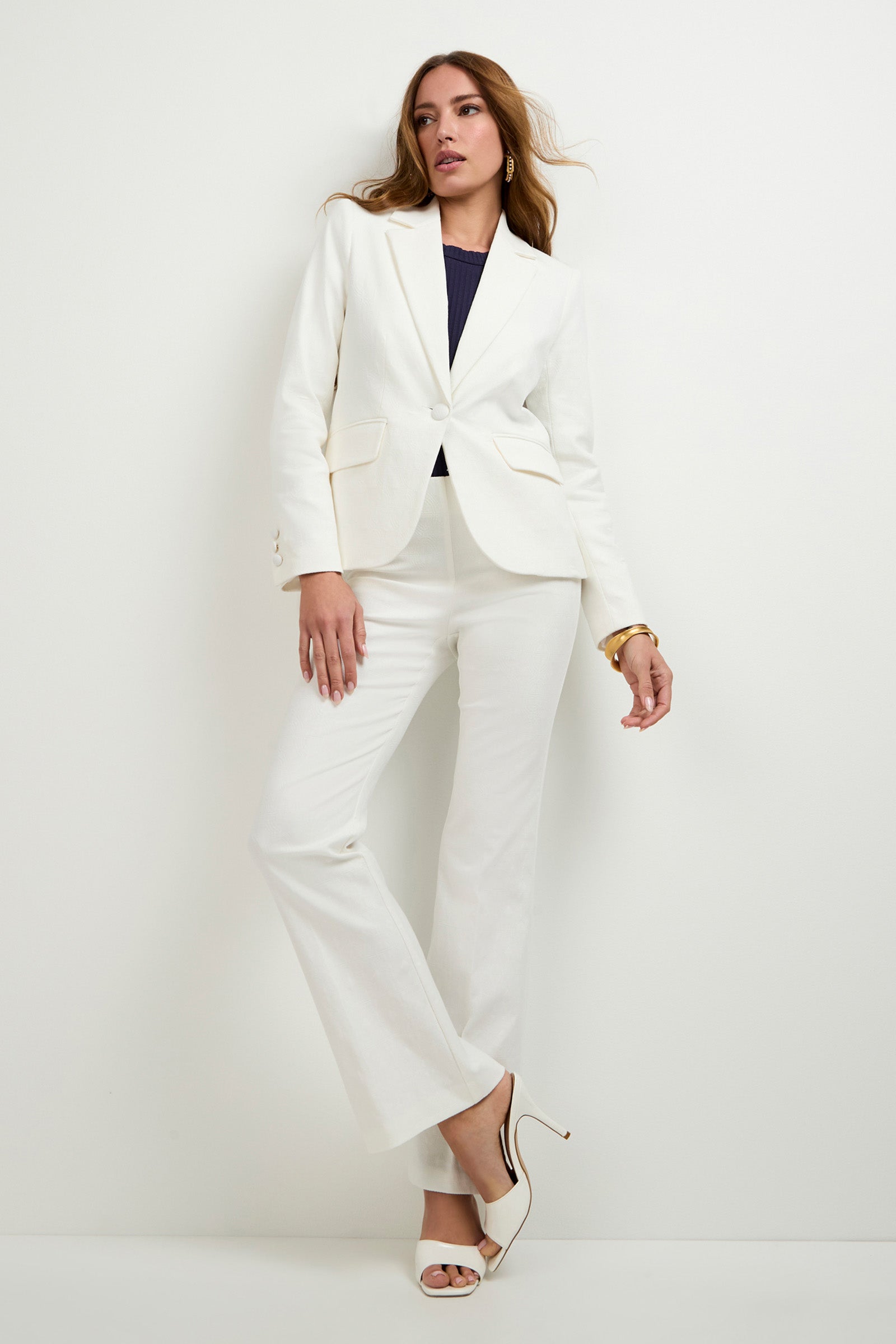 A woman stands against a white wall wearing the Incanta Fitted Blazer with wide-leg pants, a dark top, white high heels, and a gold bracelet. Her hair is down and one leg is slightly bent.