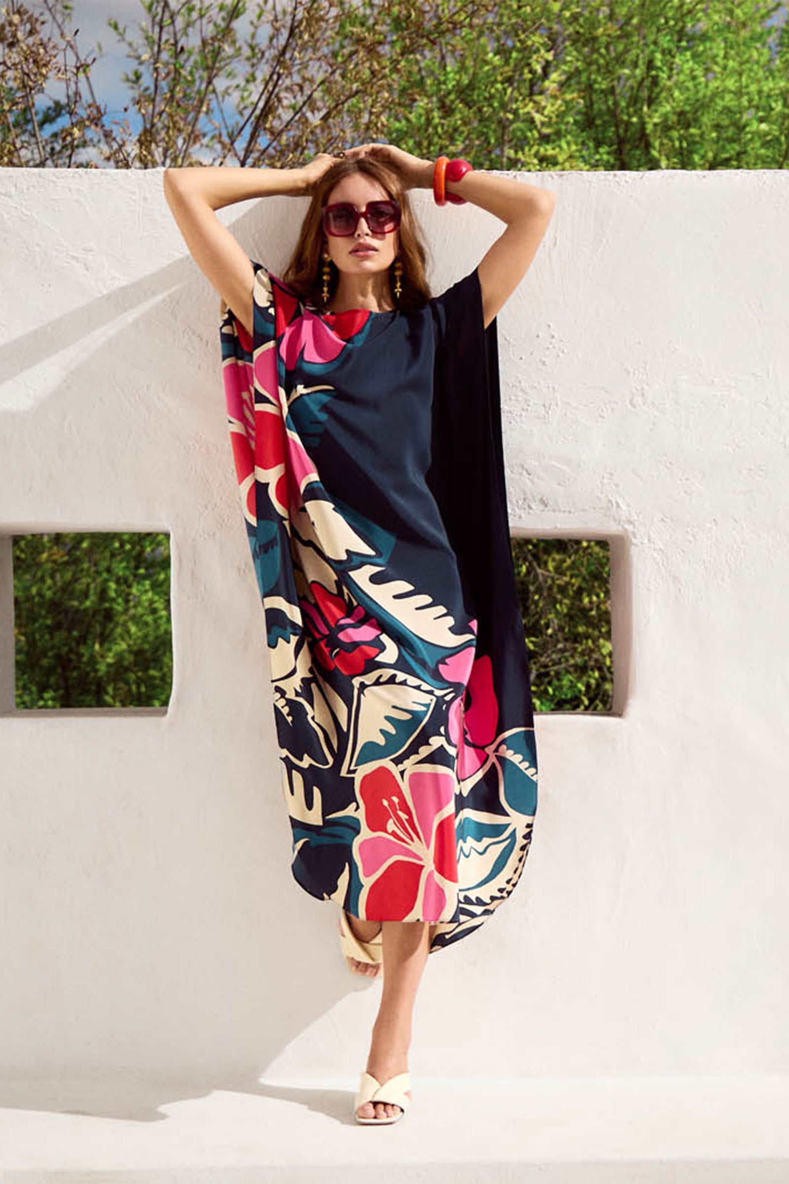 A woman poses against a white wall in the Jalani Maxi Caftan Dress, featuring a bold floral pattern in black, navy, and cream. She accessorizes with red sunglasses, a red bangle, open-toed heels, and is framed by green foliage.