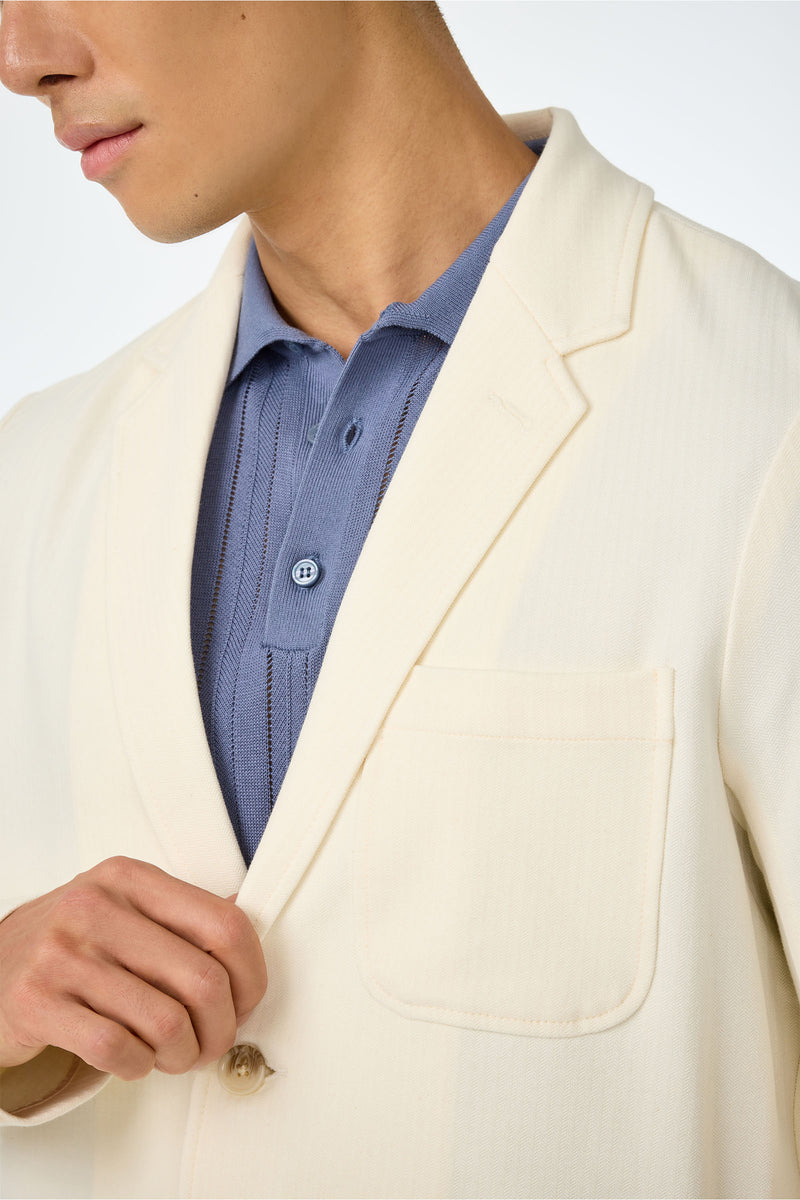 A person wearing the Muir Knit Blazer in cream over a light blue polo shirt holds its lapel with one hand. Only their lower face, neck, and upper torso are visible.