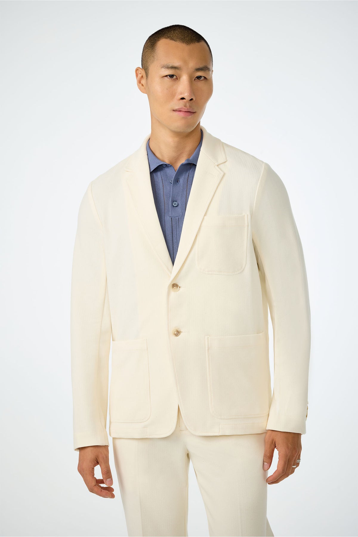 A man with short hair wears the Muir Knit Blazer in ecru over a blue collared shirt, standing against a plain light background.
