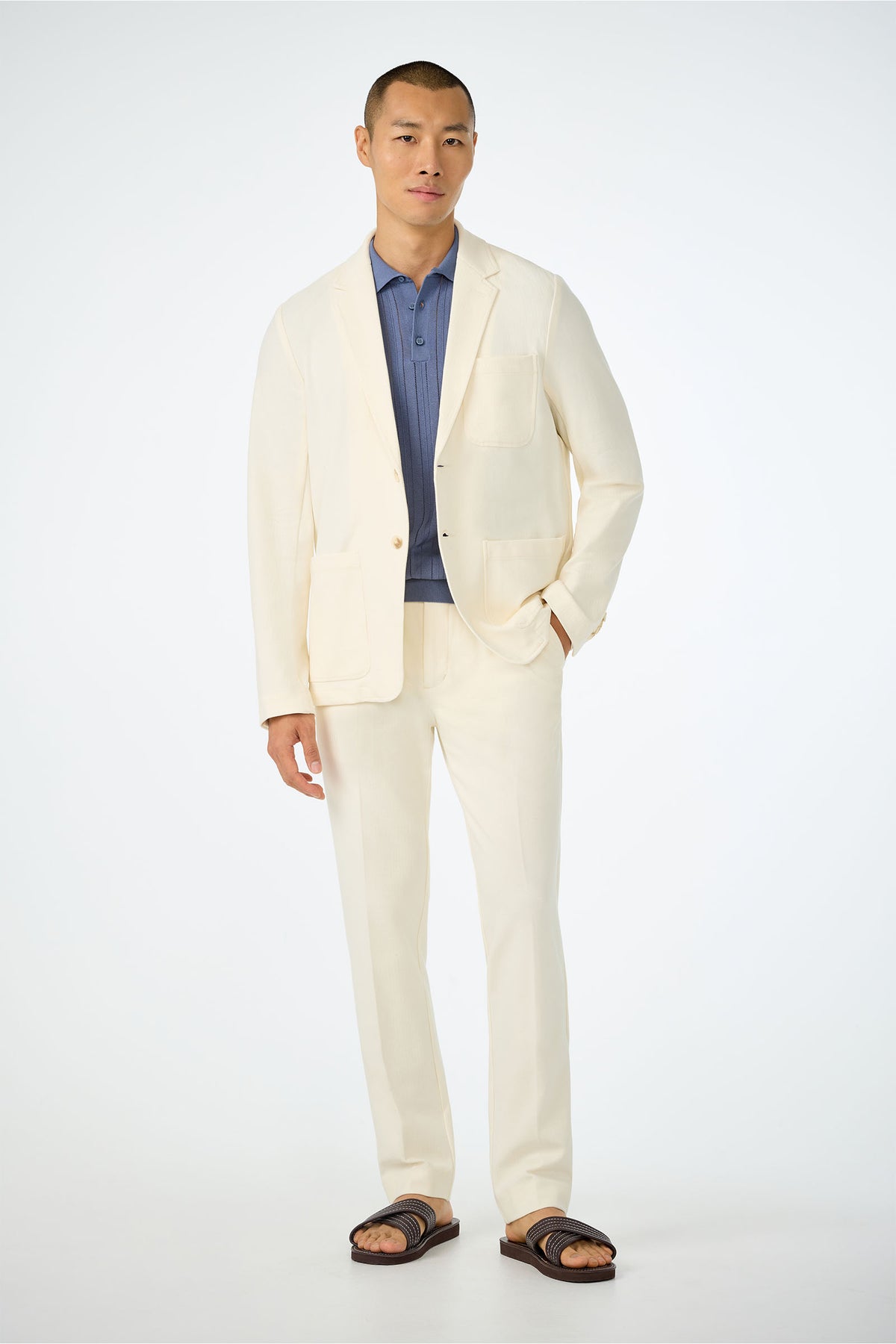 A man poses against a plain background wearing the cream-colored Muir Knit Blazer over a blue polo shirt, paired with brown sandals and with one hand in his pocket.
