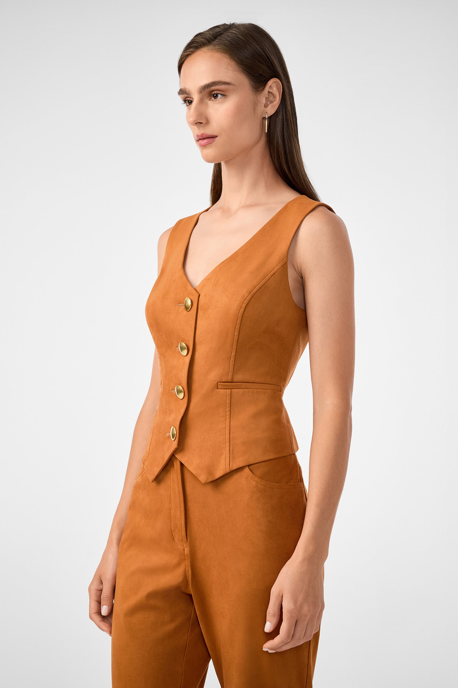 A woman with straight brown hair models the Kofi 2 Vest, a sleeveless rust-colored piece with gold buttons, paired with matching high-waisted pants against a plain light gray background, posed slightly to the side.
