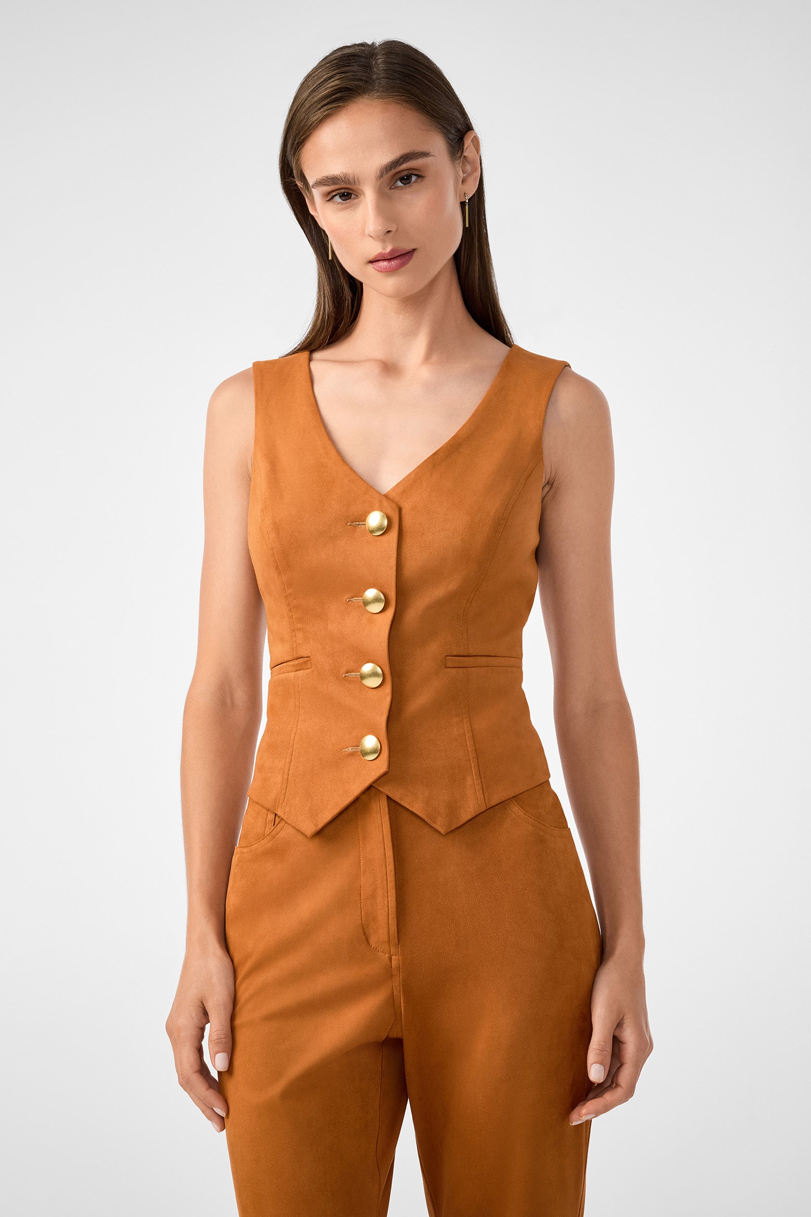 A woman with straight brown hair wears the sleeveless, burnt orange Kofi 2 Vest with gold buttons and matching high-waisted pants, posing against a plain light gray background.
