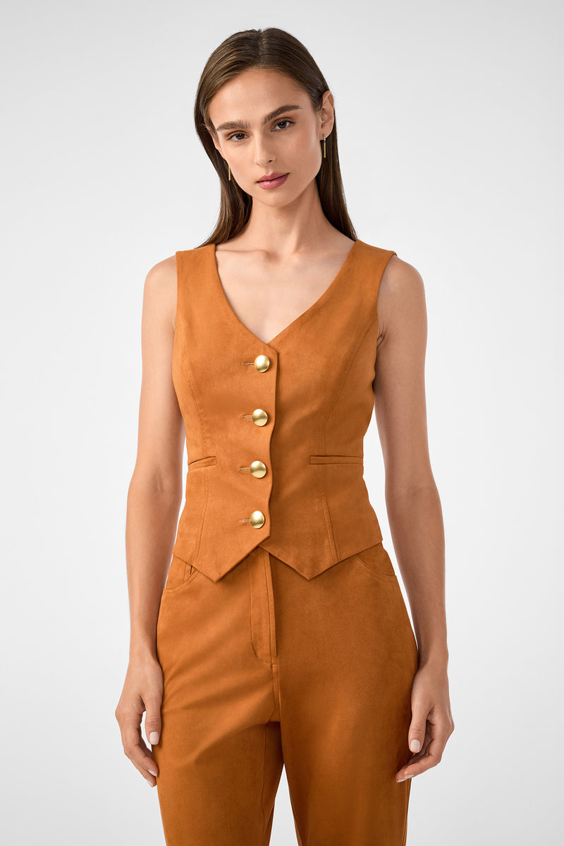 A woman with straight brown hair wears the sleeveless, burnt orange Kofi 2 Vest with gold buttons and matching high-waisted pants, posing against a plain light gray background.
