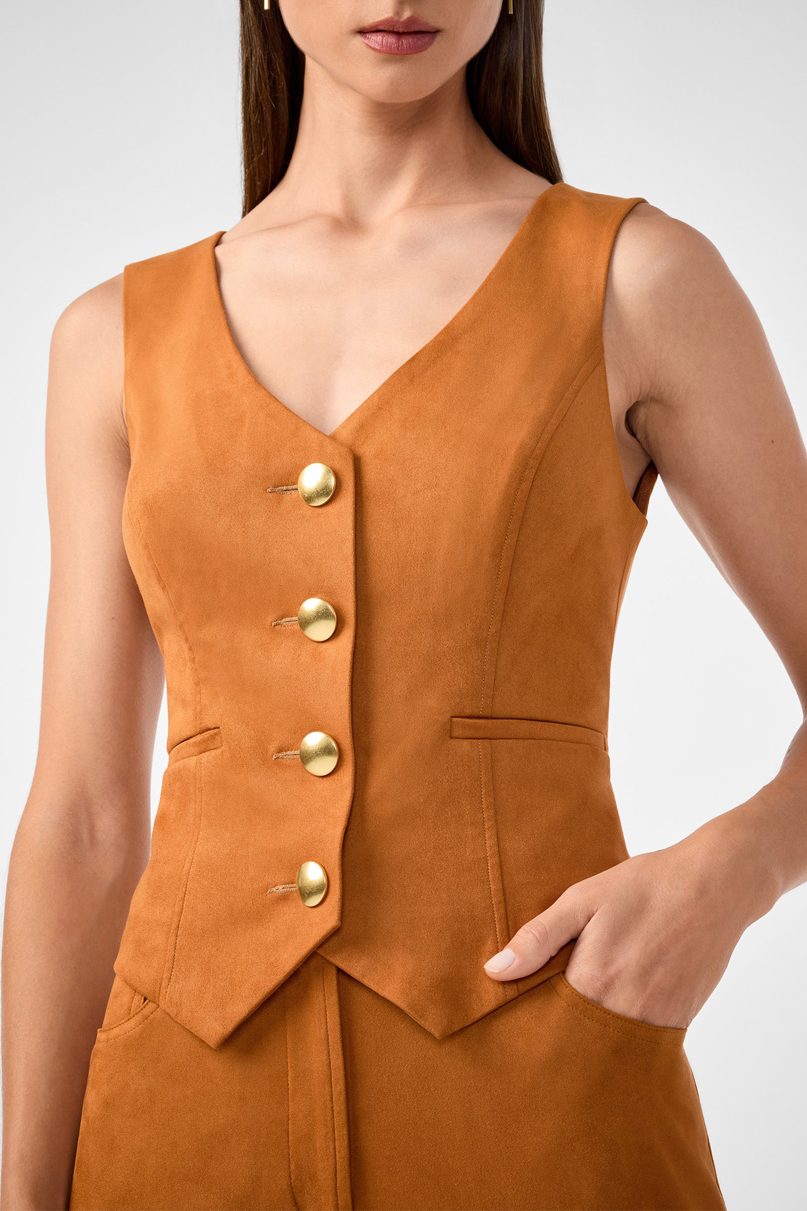 A woman wears the Kofi 2 Vest, a sleeveless tan suede vest with large gold buttons, V-neckline, and pointed hem. One hand is in her pocket; image cropped at shoulders and mid-thigh.