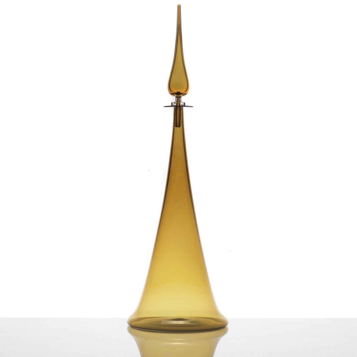 The Large Fluted Cone Decanter, a tall, slender amber glass bottle with a long, pointed stopper, is displayed on a reflective white surface against a plain white background.