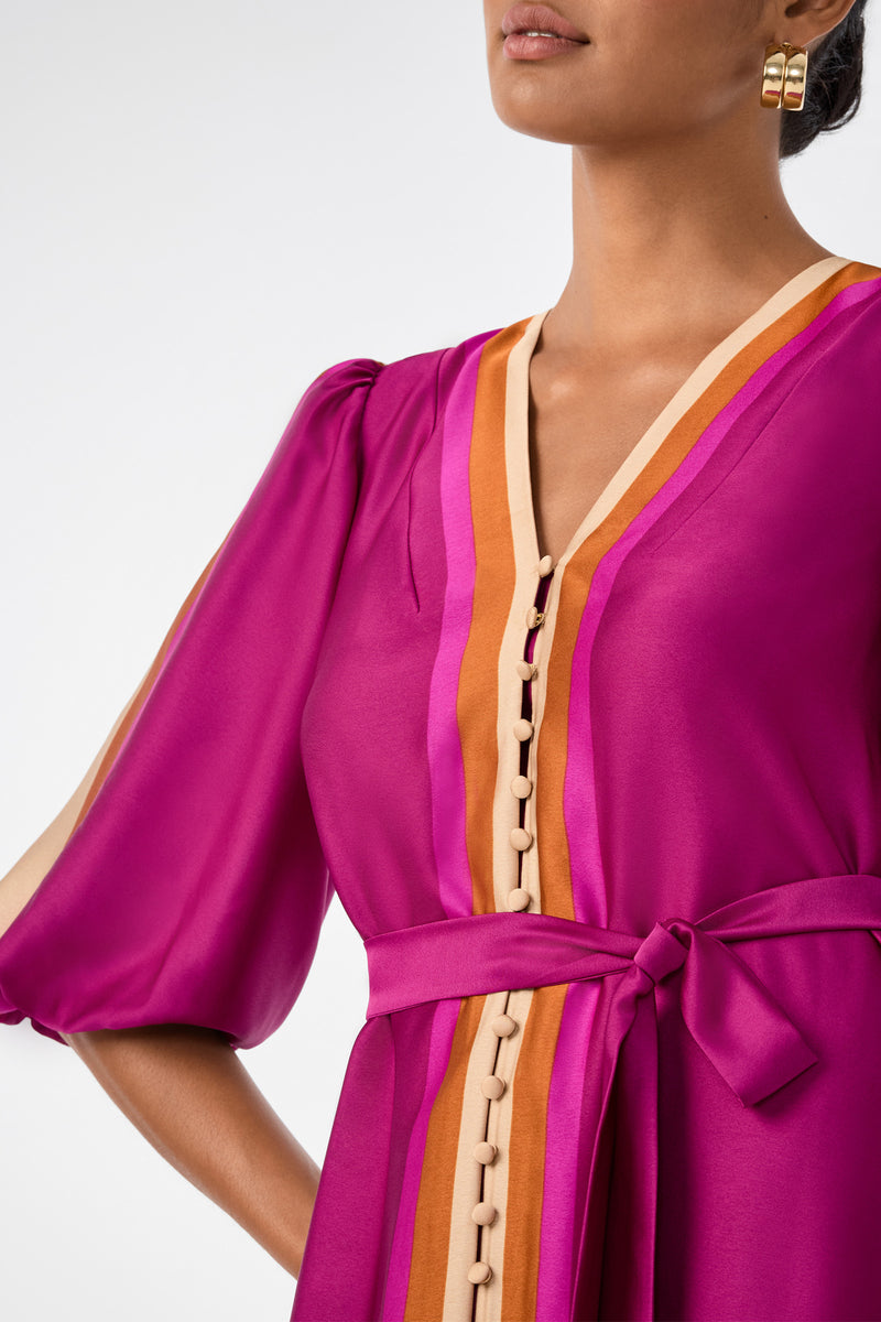 A woman wears the Linden Dress, a vibrant magenta style with orange and beige trim, button details, and a matching waist tie. She accessorizes with gold hoop earrings and her hair pulled back. Image is cropped at her nose and above the waist.