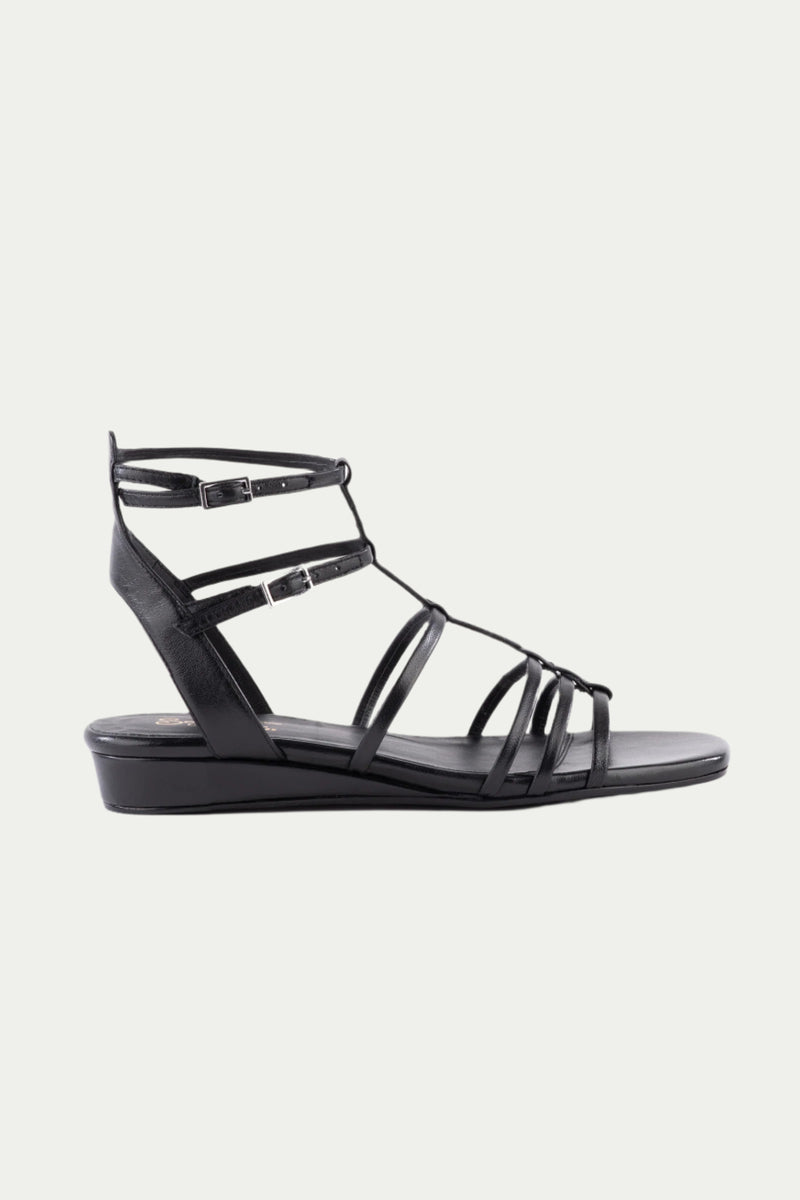 The Luxurious Wedge Sandal - Black Leather features black straps, a low wedge heel, ankle buckles, and an open toe, showcased in side profile against a plain white background.