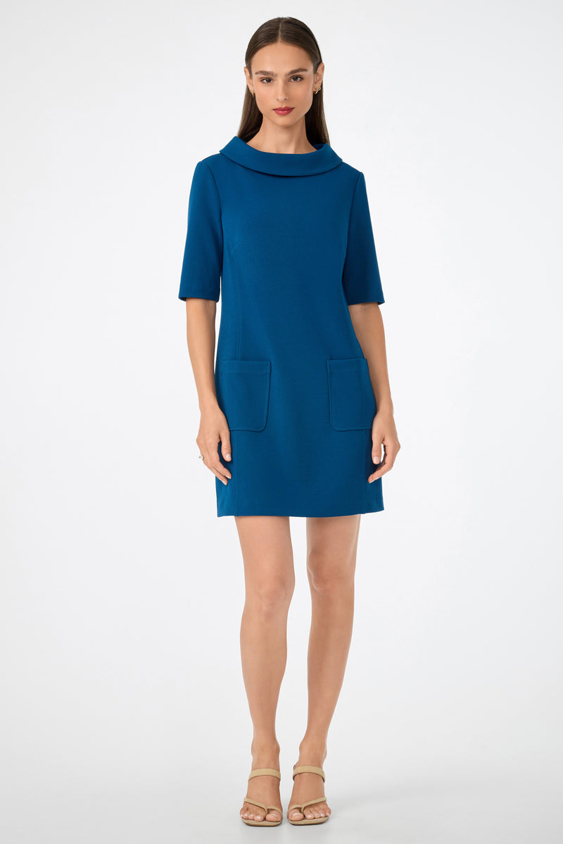 A woman faces forward wearing the Maleko Dress—a teal, knee-length style with short sleeves, a wide collar, and two front pockets. She has straight brown hair and beige heeled sandals, standing against a plain light background.
