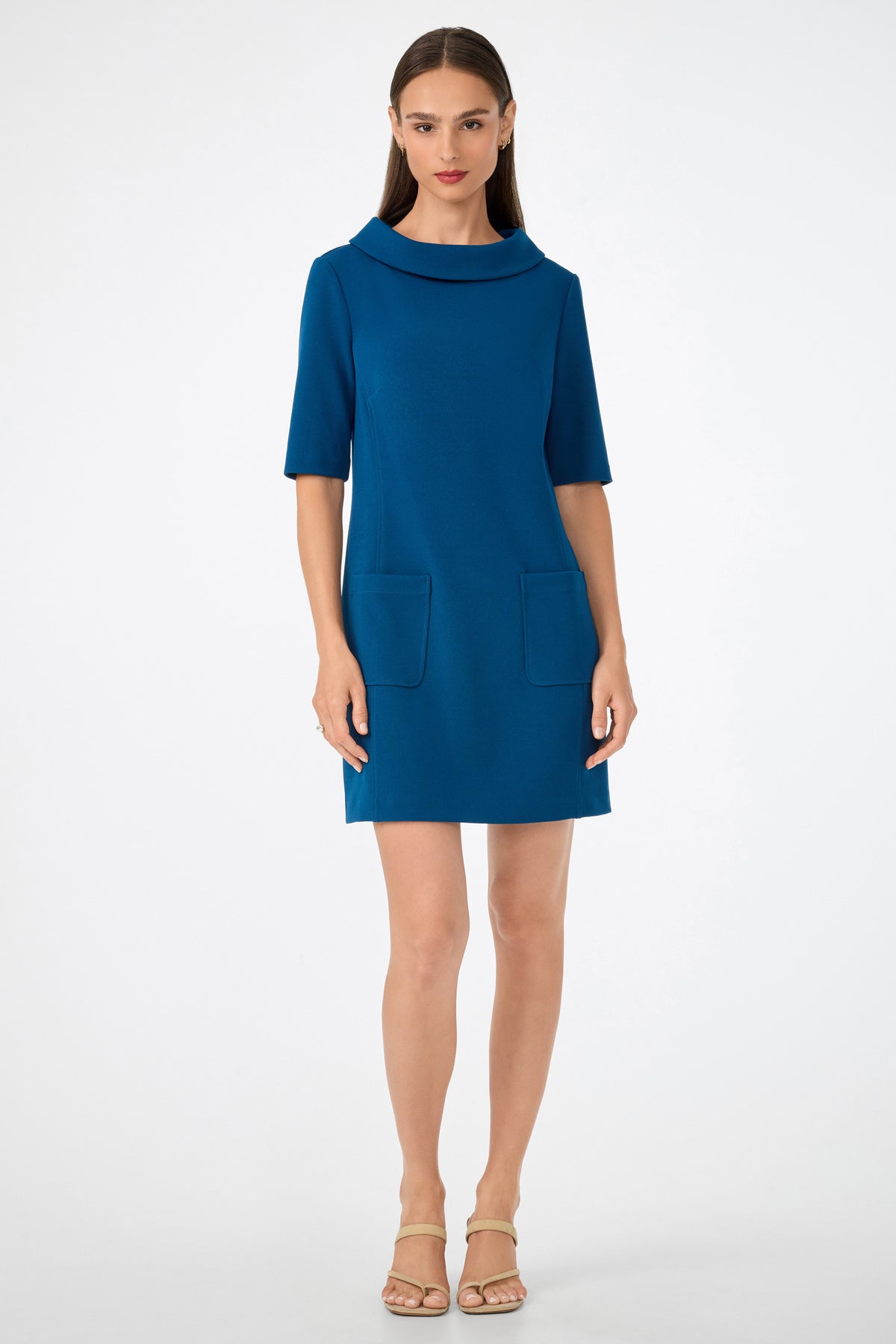 A woman faces forward wearing the Maleko Dress—a teal, knee-length style with short sleeves, a wide collar, and two front pockets. She has straight brown hair and beige heeled sandals, standing against a plain light background.
