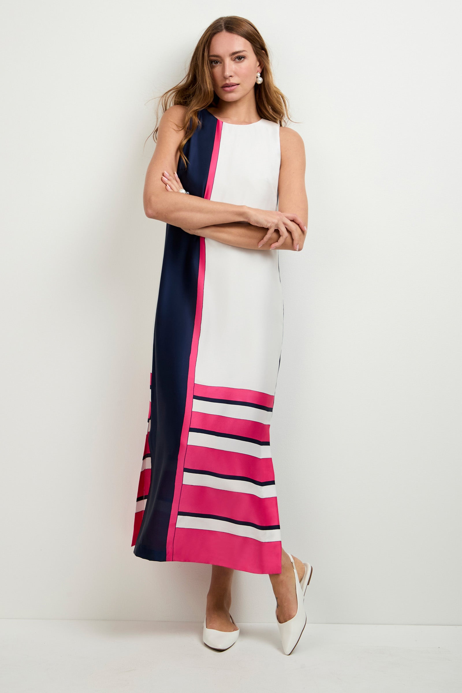 A woman poses against a white wall wearing the Moderna Linee Midi Dress, a sleeveless, ankle-length piece with white, navy blue, and pink color-blocked stripes, paired with white pointed-toe shoes.
