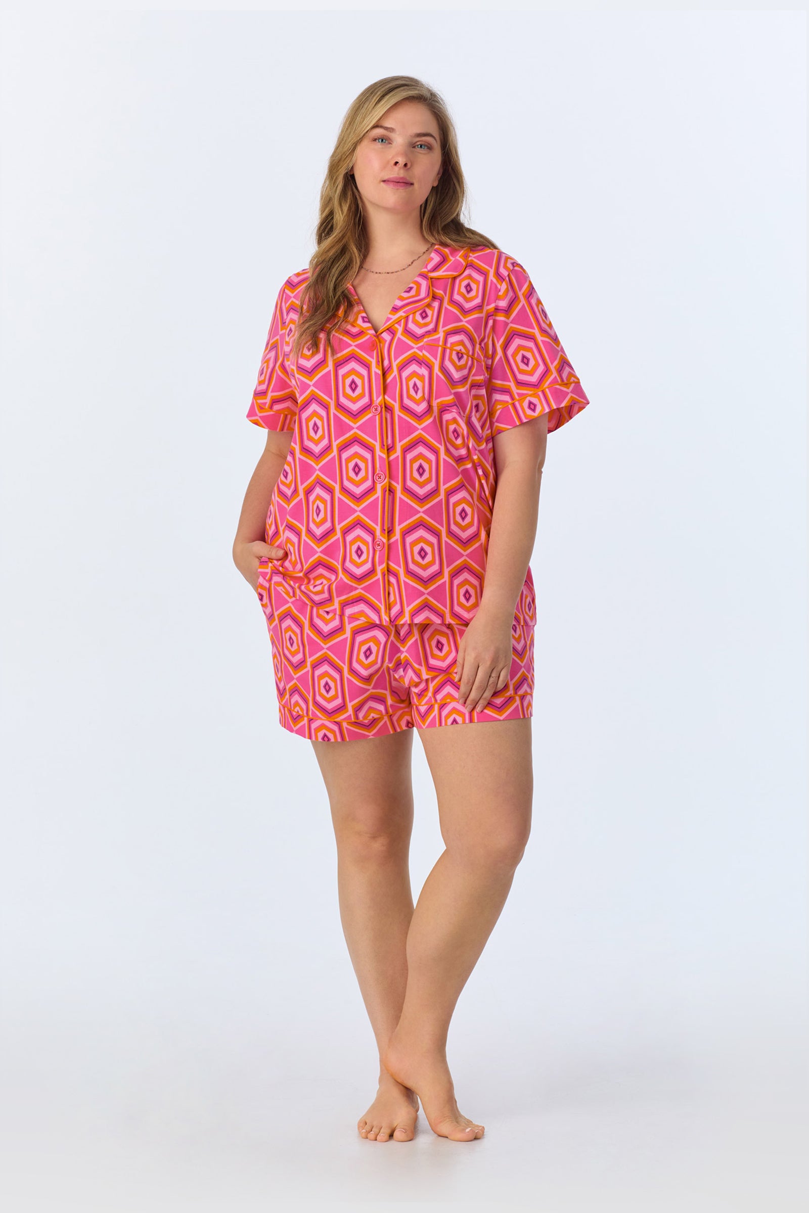 A woman stands barefoot against a light background, wearing the TT SS Shorty PJ Set - MLT Hexagon Jersey, a short-sleeved, button-up romper with a bold pink, orange, and white geometric pattern. She looks relaxed with one hand in her pocket.