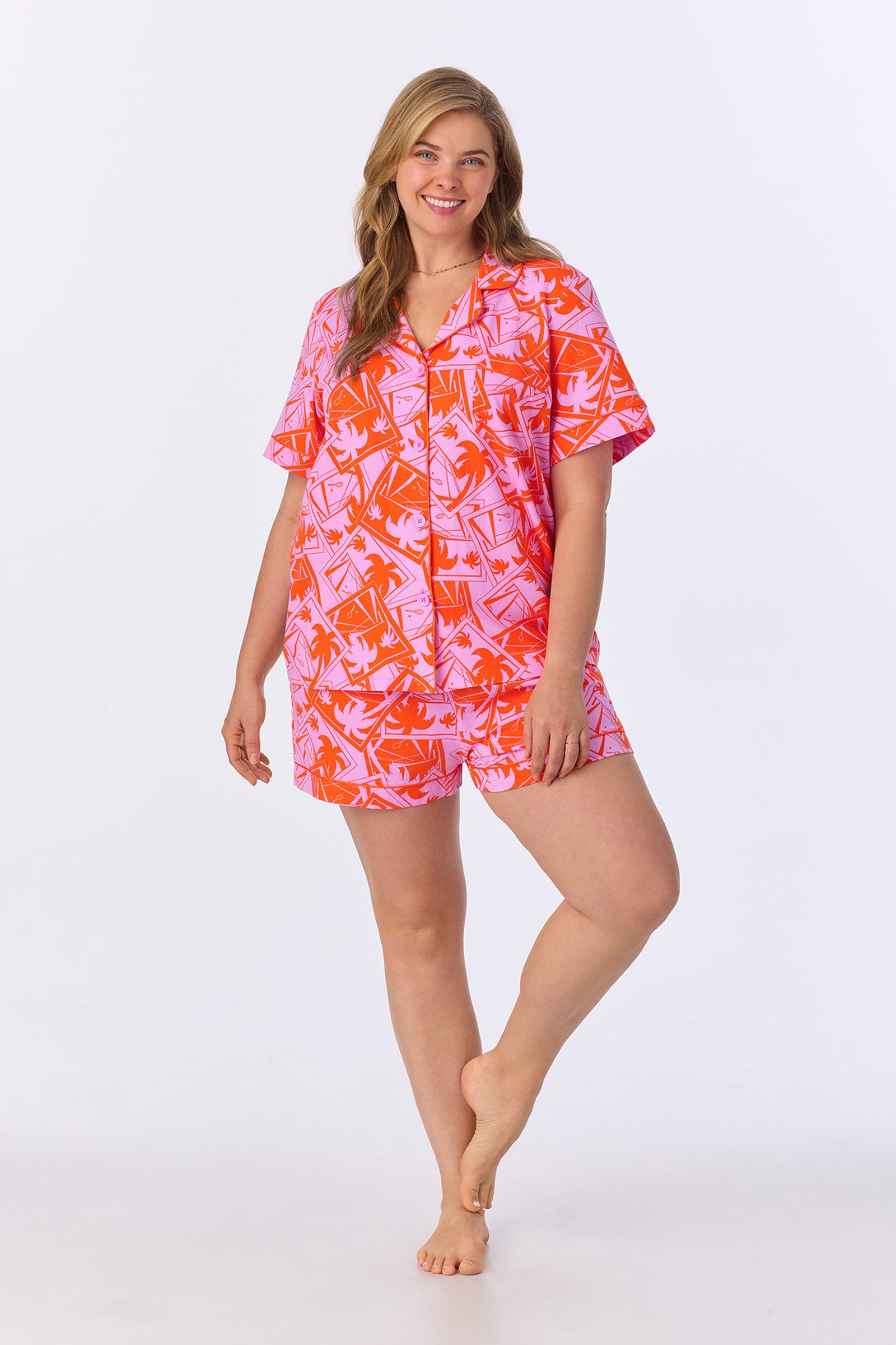 A smiling woman with light brown hair models the Shorty PJ Set - Tennis Stretch Jersey, featuring a colorful pink and orange short-sleeve button-up shirt and matching shorts, standing barefoot against a plain white background.