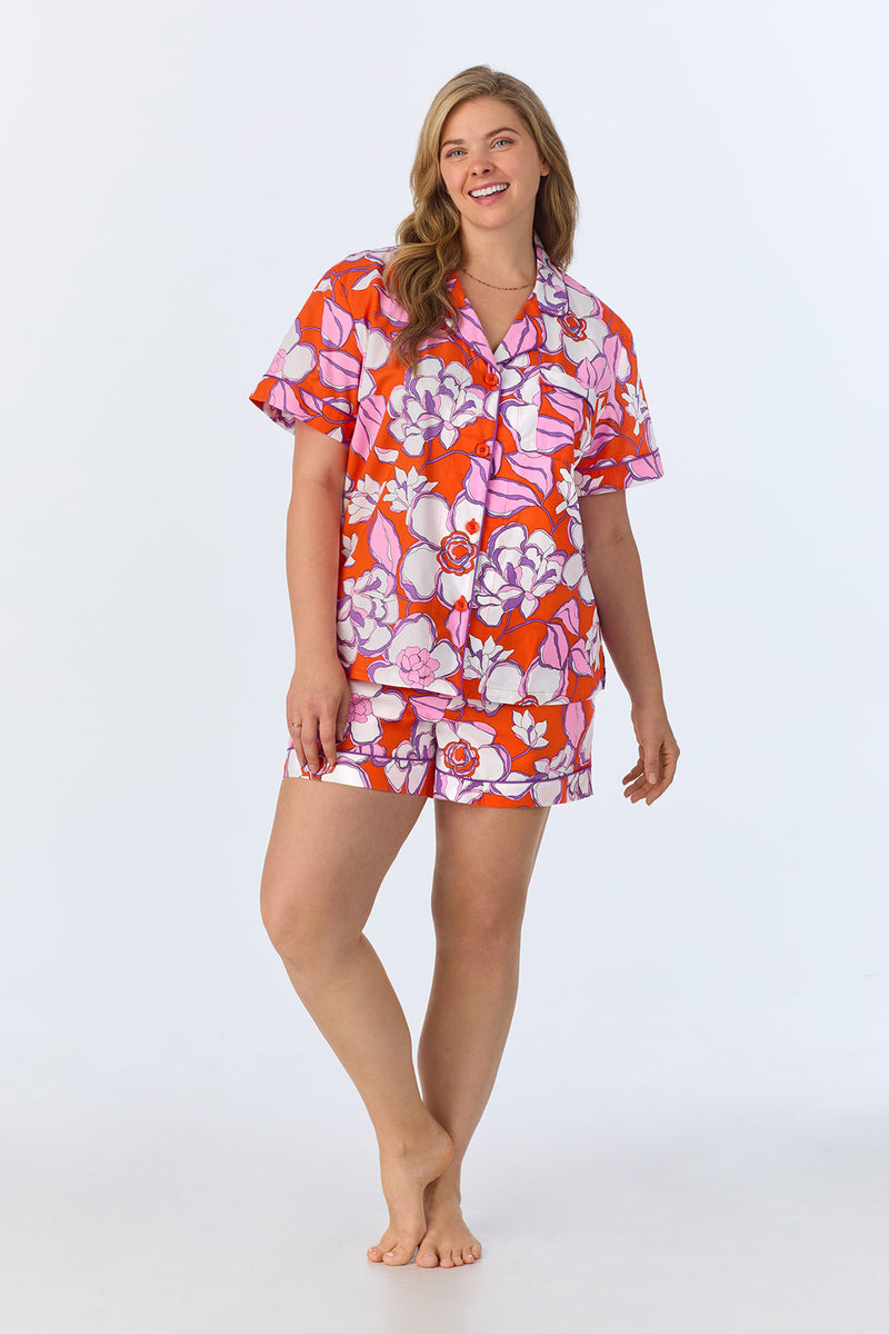 A woman with long, light brown hair stands barefoot and smiling, wearing the Shorty PJ Set - Gardenia Poplin: a colorful floral short-sleeve button-up top and matching shorts in red, pink, and white on a plain light background.