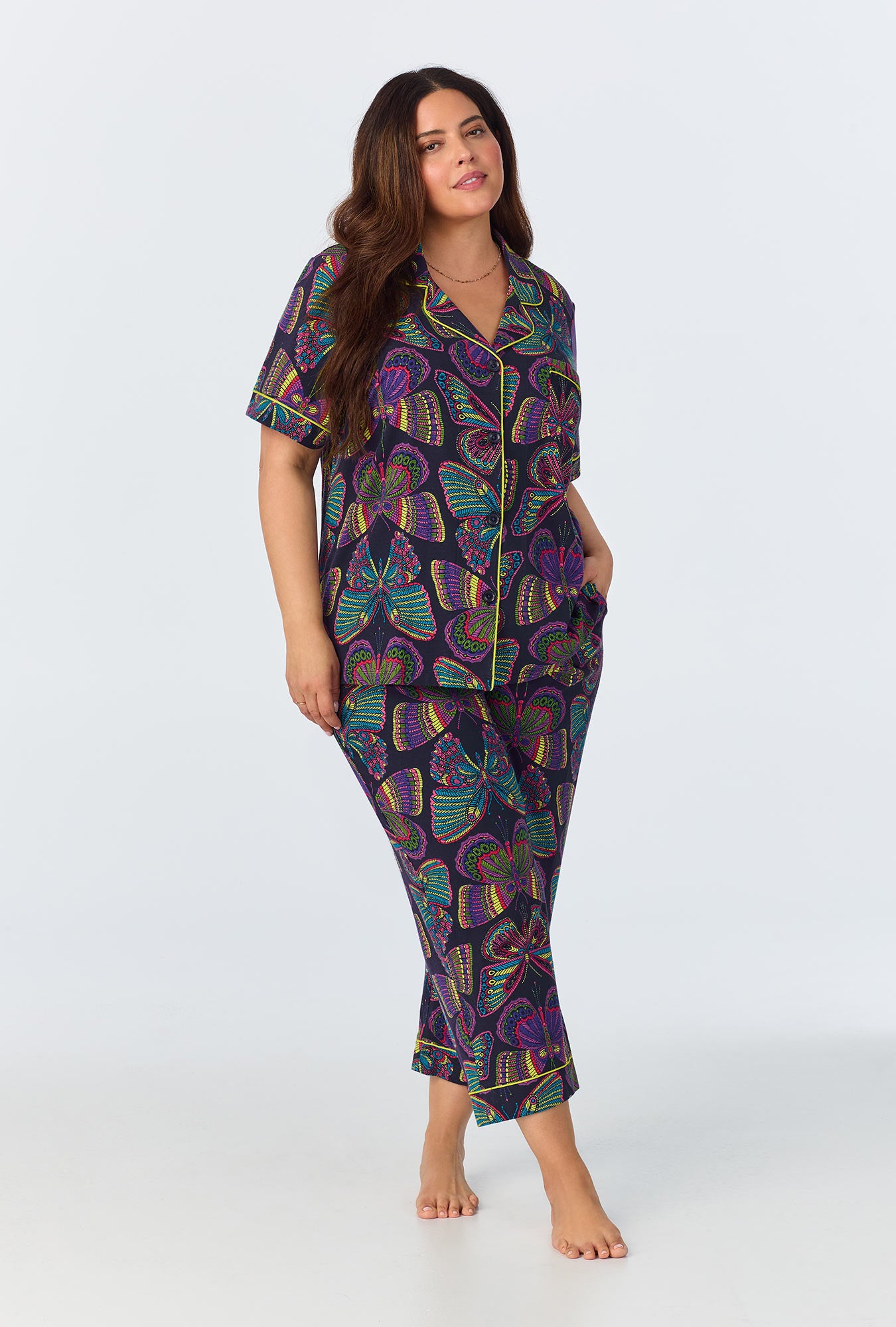 A woman with long dark hair wears the Butterflies Womens Short Sleeve Cropped Pant Jersey PJ Set in Multi, featuring a colorful abstract design. The PJ set includes a short-sleeve button-up top and cropped pants. She stands barefoot against a plain light background.