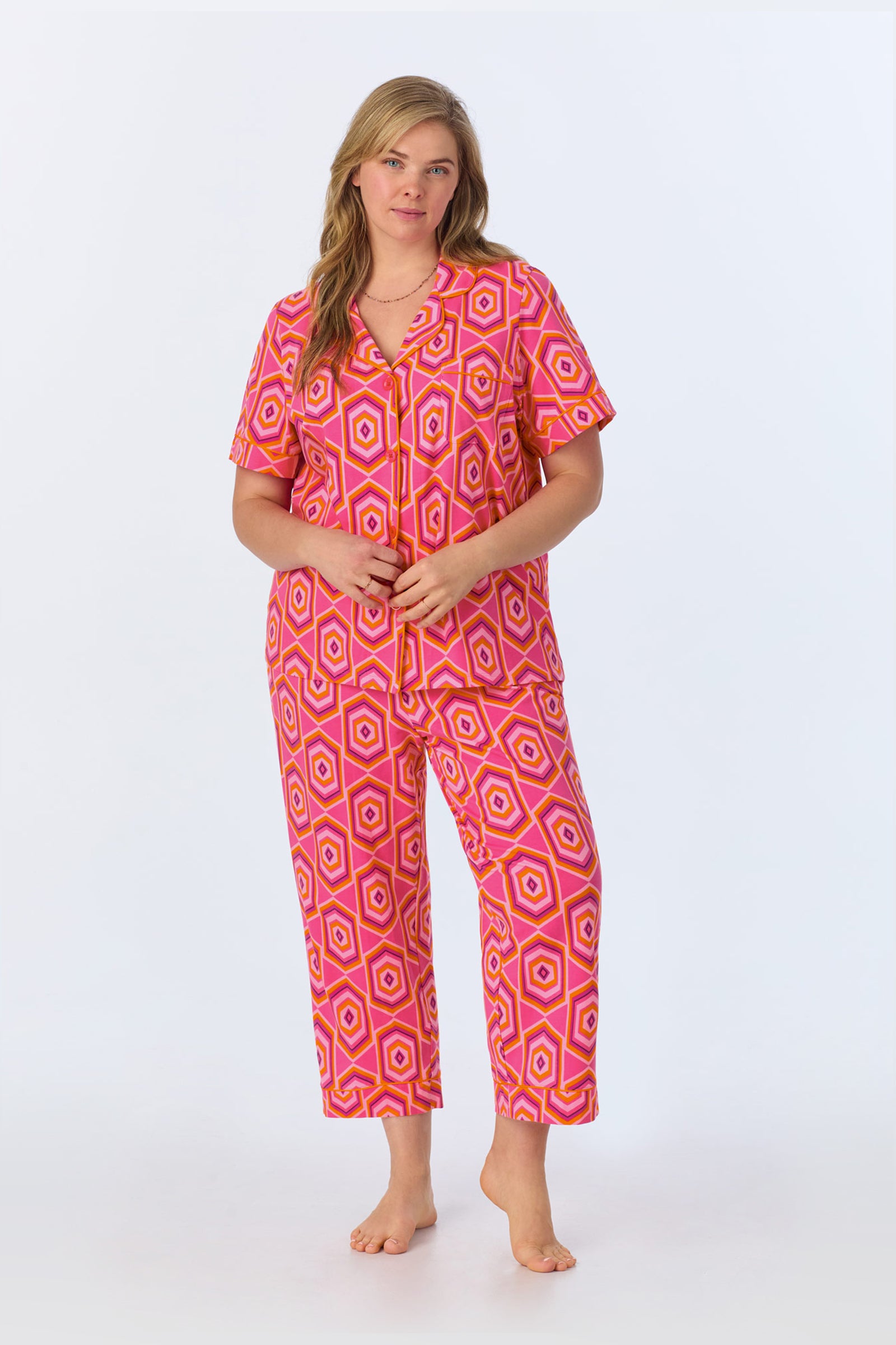 A woman stands barefoot, wearing the TT SS Crop PJ Set - MLT Hexagon Jersey, a short-sleeve button-up jumpsuit with a bright pink, orange, and white geometric hexagon pattern against a plain white background.