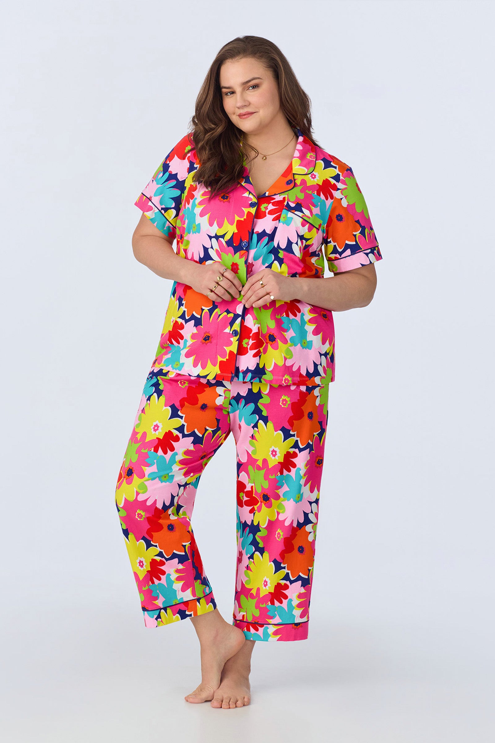 A woman stands barefoot in the Crop Classic PJ Set - Pop Floral Stretch Jersey, featuring a vibrant pink, yellow, green, orange, and blue floral pattern. The set includes short sleeves and cropped pants as she poses confidently against a plain background.