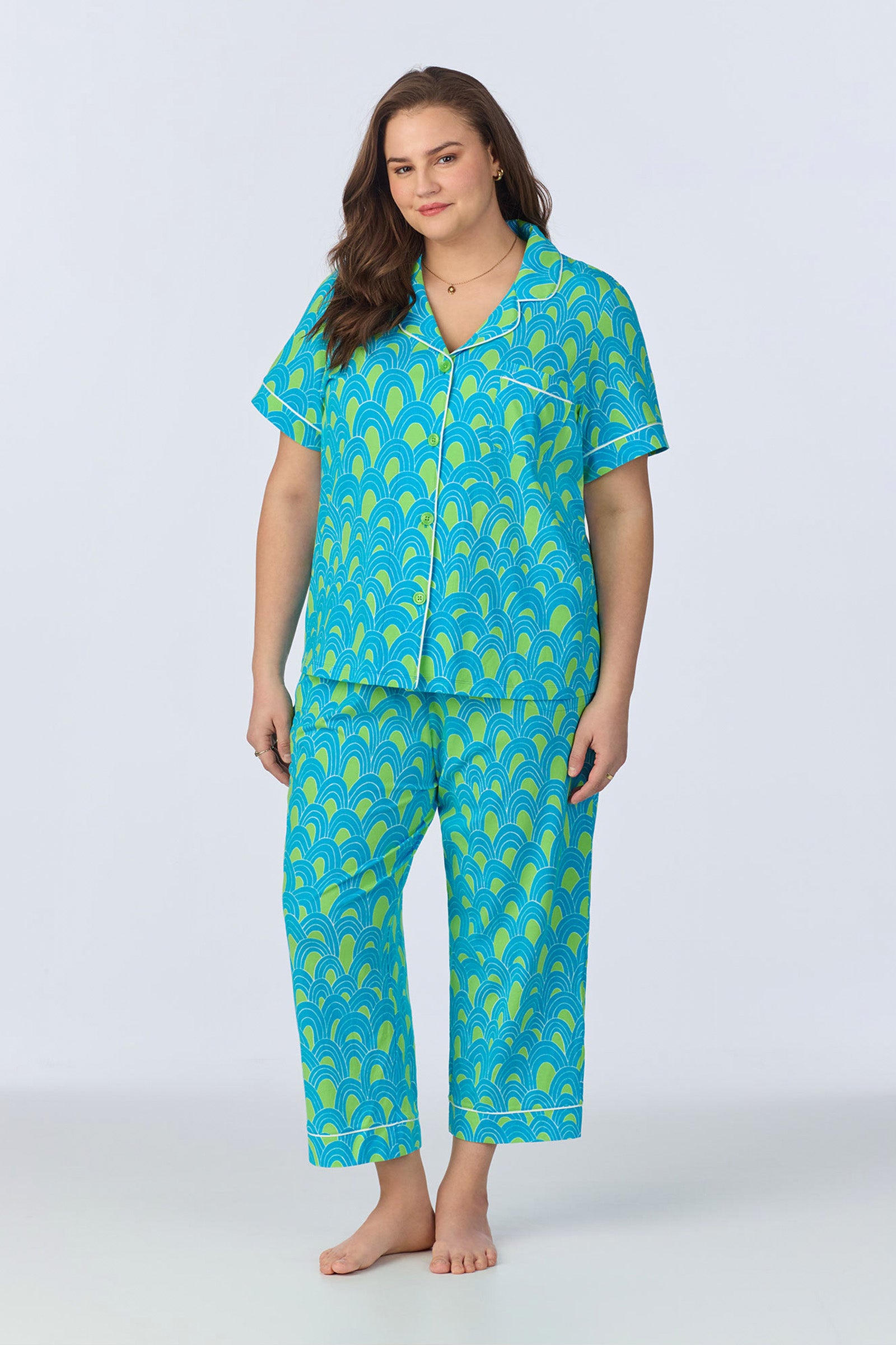 A woman stands barefoot, smiling gently in the Crop Classic PJ Set - Geo Stretch Jersey. The bright blue set features a bold yellow and green scallop design, short sleeves, button-up top, and long pants against a plain light background.