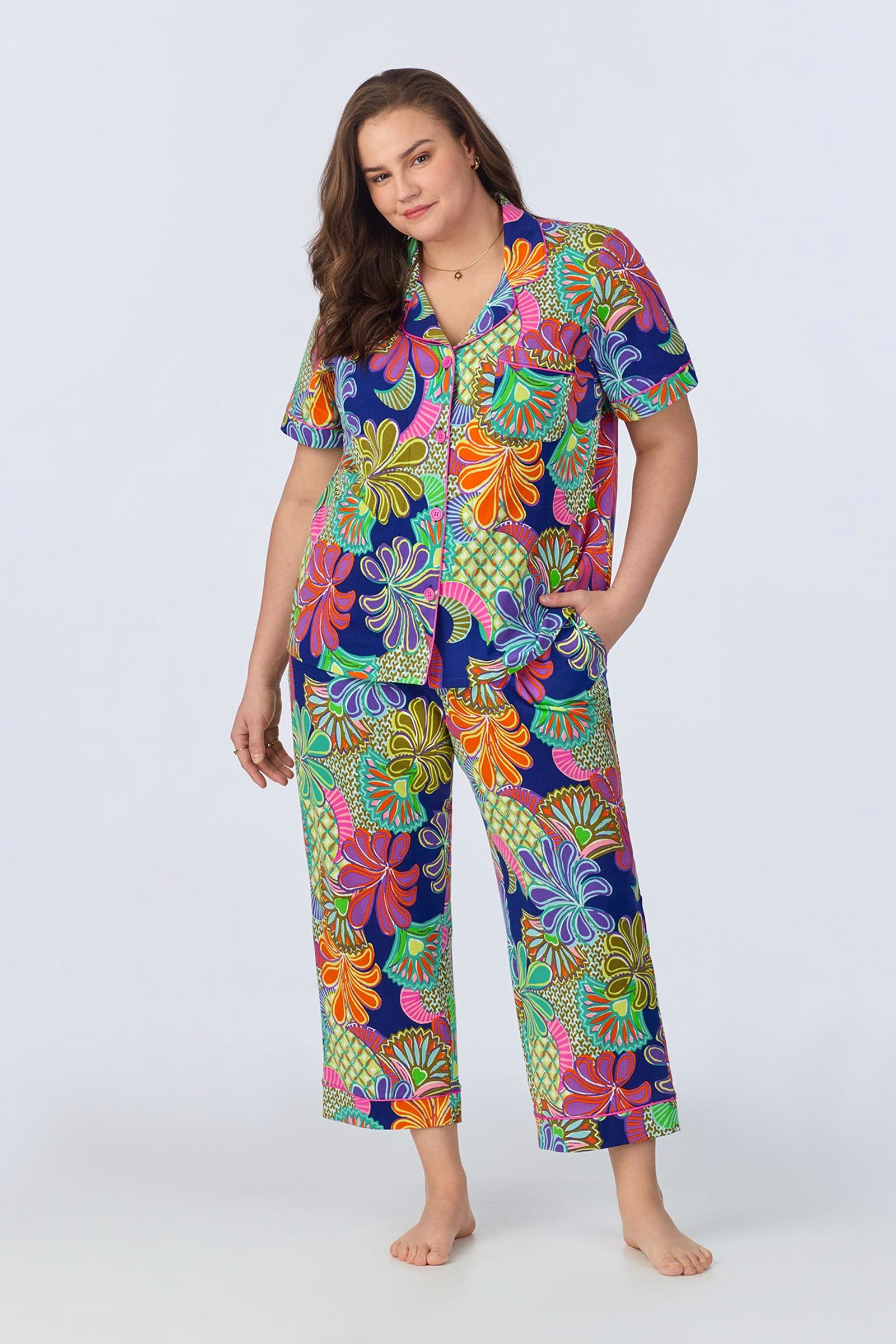 A woman with long brown hair wears the Crop Classic PJ Set - Zanzibar Stretch Jersey, featuring a colorful floral short-sleeve button-up top and matching pants. She stands barefoot, one hand in her pocket, against a light background.