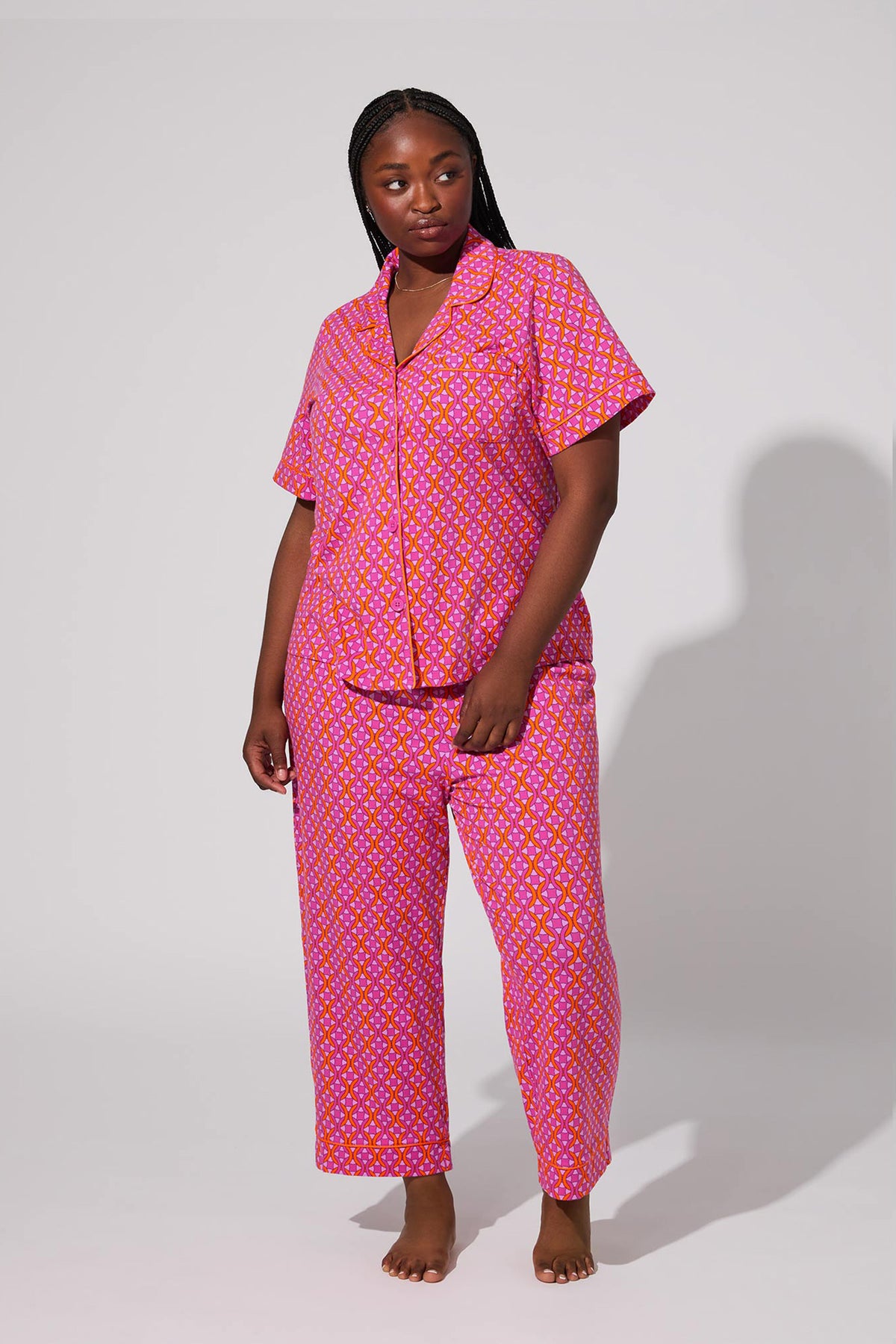 A woman stands barefoot against a light gray background, wearing the Crop Classic PJ Set - Chain Geo, featuring a matching short-sleeve collared button-down top and long pants in a stylish chain geo pattern.