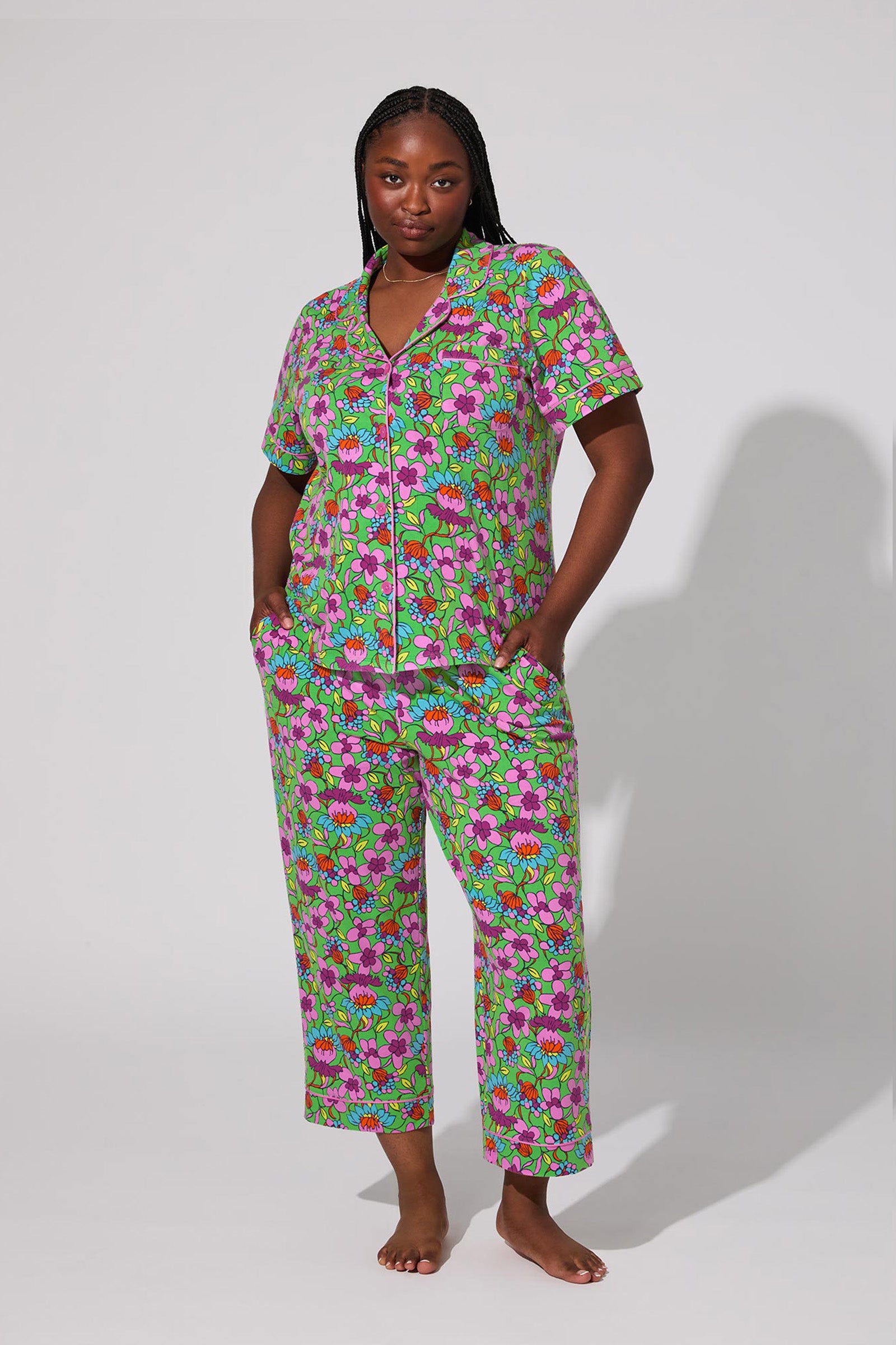 A woman stands barefoot against a plain background, wearing the Crop Classic PJ Set - Garden Floral, a matching short-sleeve pajama set with a vibrant green, purple, pink, and blue floral print. She places her hands in her pockets and looks confidently at the camera.