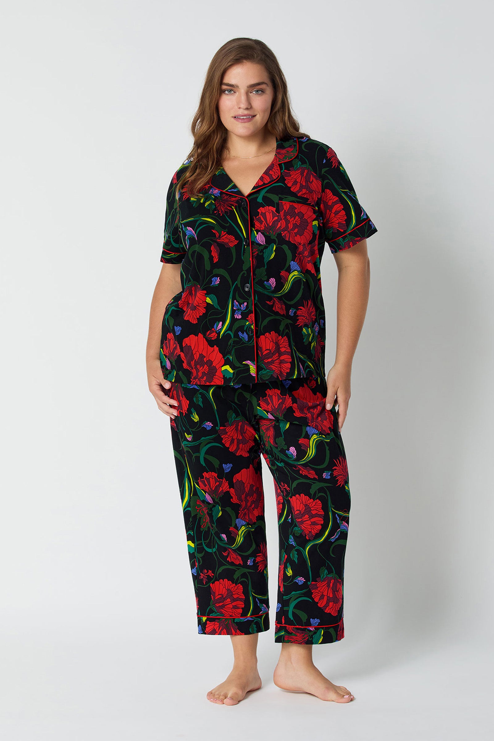 A woman with long brown hair wears the "Haute Floral S/S Cropped Pj Set," featuring a bold red floral pattern with green leaves on black. She stands barefoot against a light gray backdrop, smiling gently.