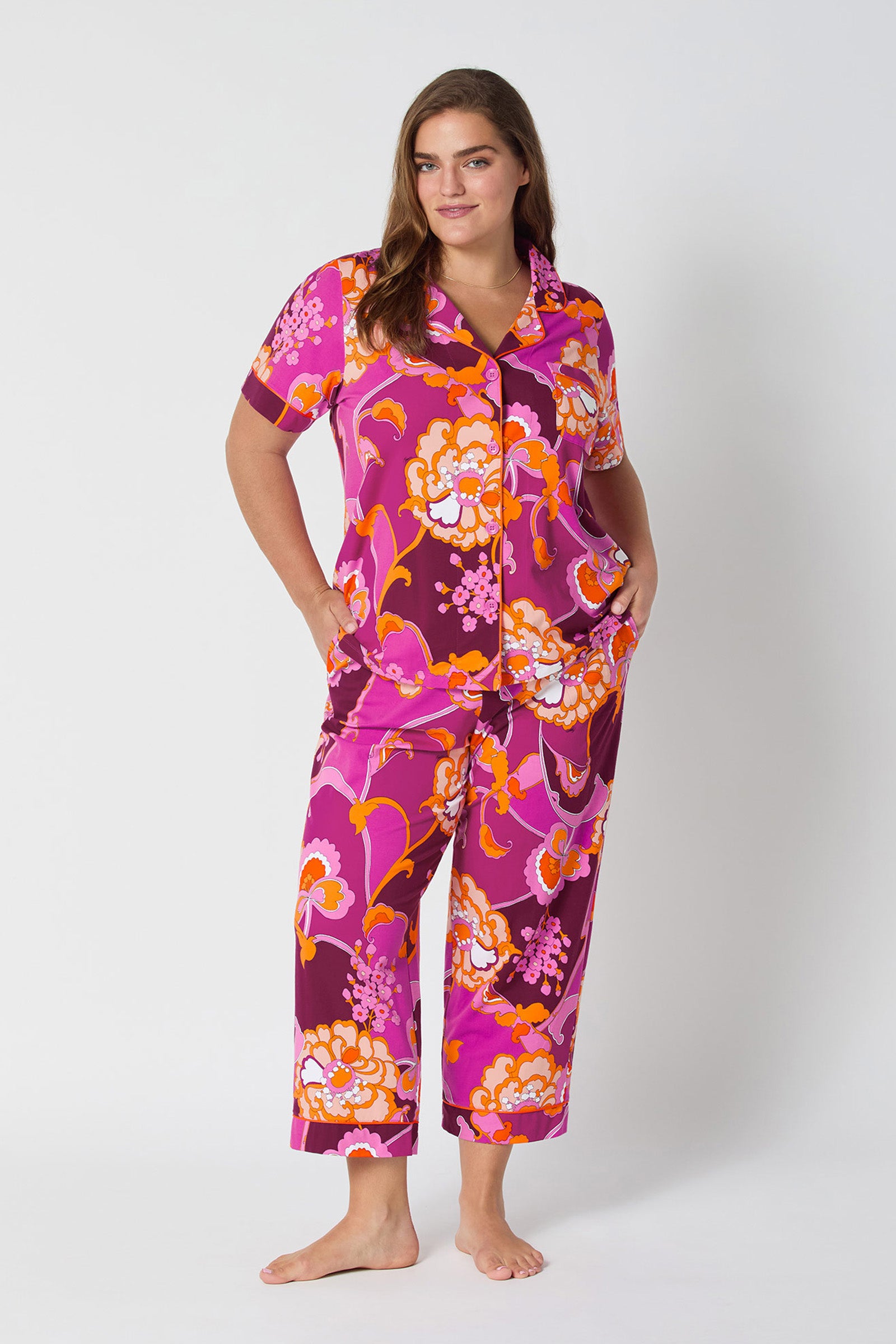 A woman stands barefoot against a white background, wearing the Apache Plume S/S Cropped PJ—vibrant magenta, orange, and white floral-print pajamas featuring short sleeves and cropped pants. She smiles slightly with her hands in her pockets.