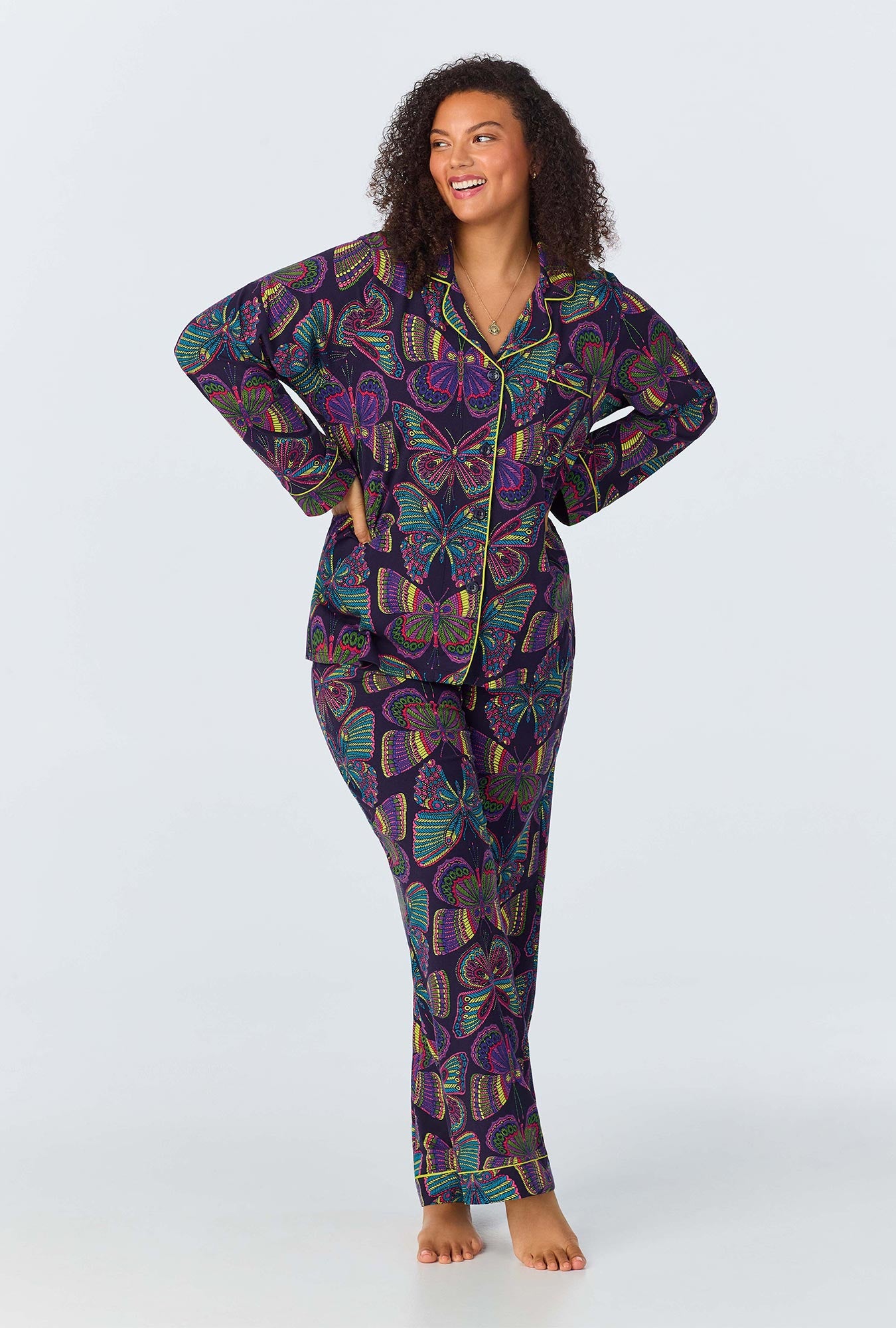 A woman stands barefoot in the Butterflies Womens Long Sleeve Long Pant Jersey PJ Set in Multi, smiling and looking to the side with hands on hips, against a plain light background.