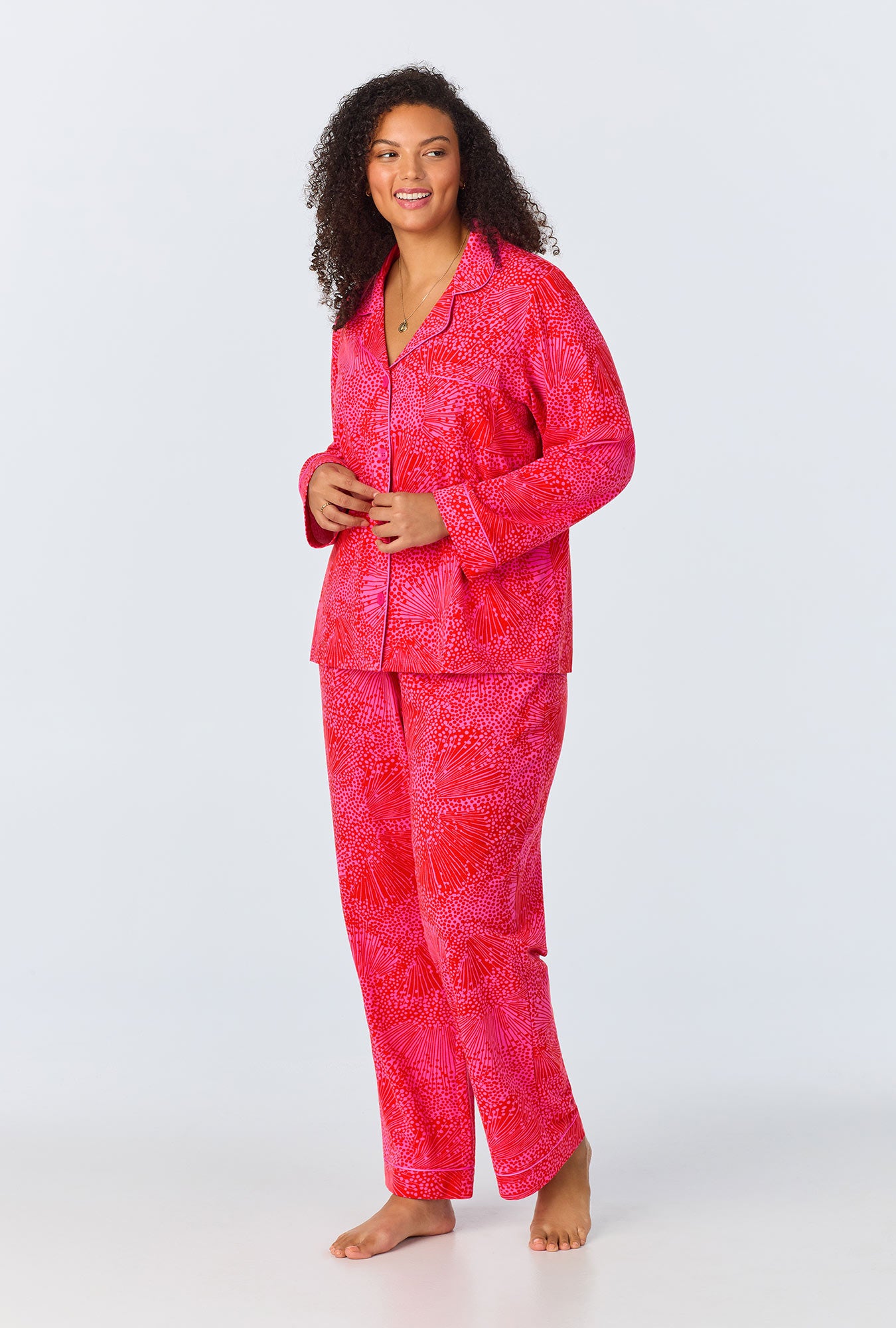 A woman stands barefoot against a white background, smiling and looking to the side in the Starburst Womens Long-Sleeve Long Pant Jersey PJ Set in Multi, featuring chic button-up top and matching pants that highlight stylish women’s sleepwear.